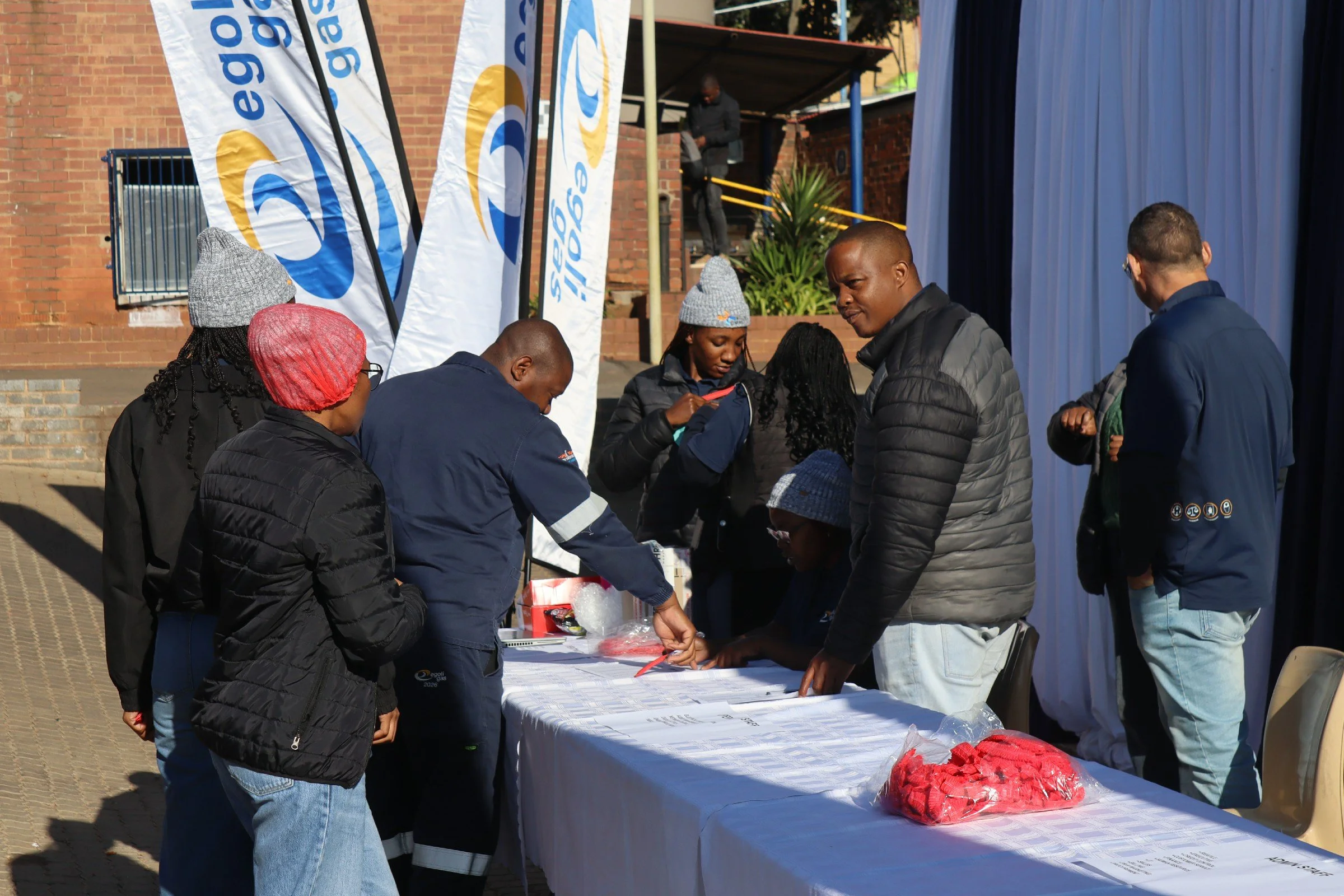People gathered around a table at an outdoor event, with flags displaying the logo of an organization, possibly for registration or distribution.