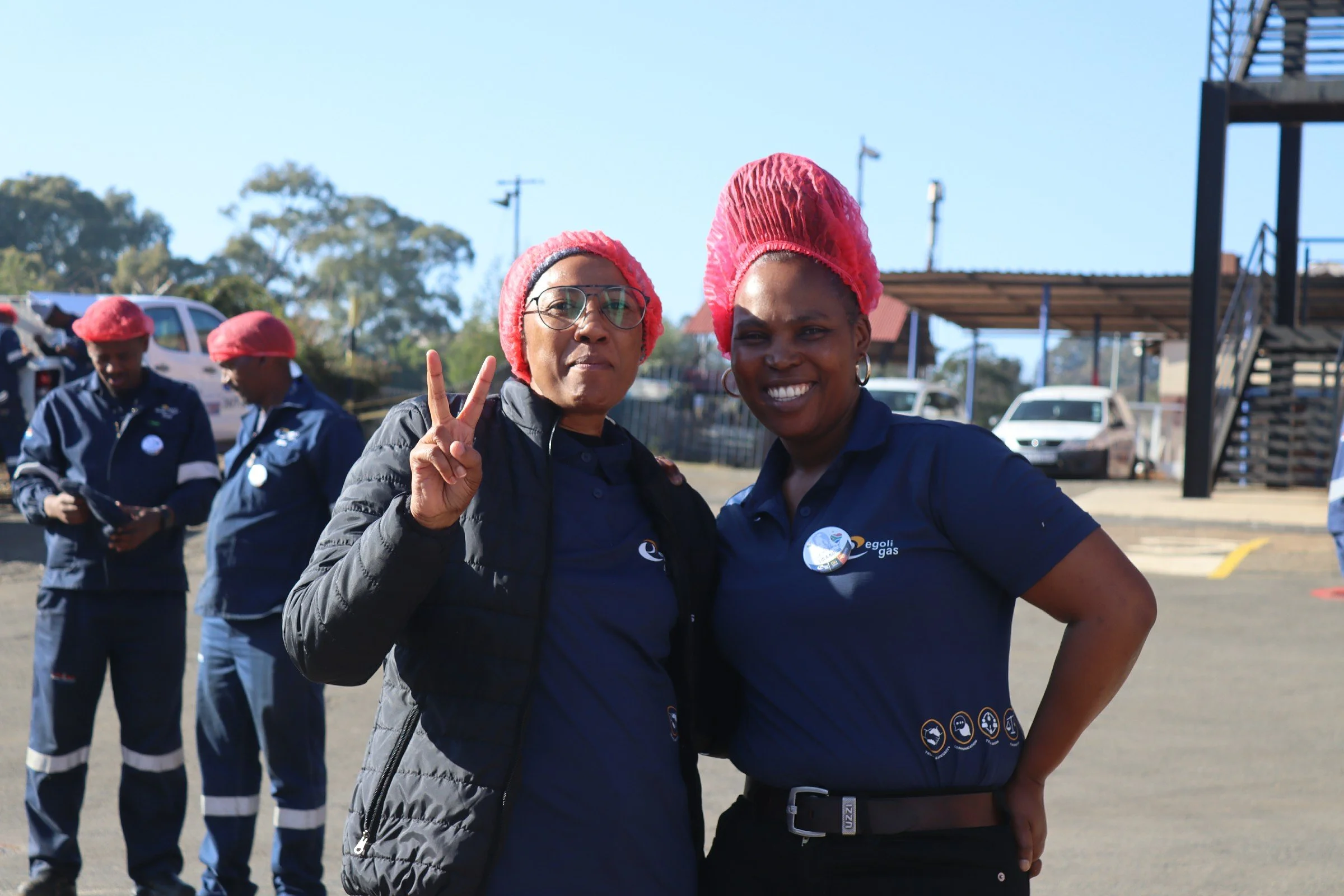 Two women wearing black outfits and pink hairnets posing outdoors, making peace signs and smiling; background includes other people in uniforms and safety gear, vehicles, and trees.