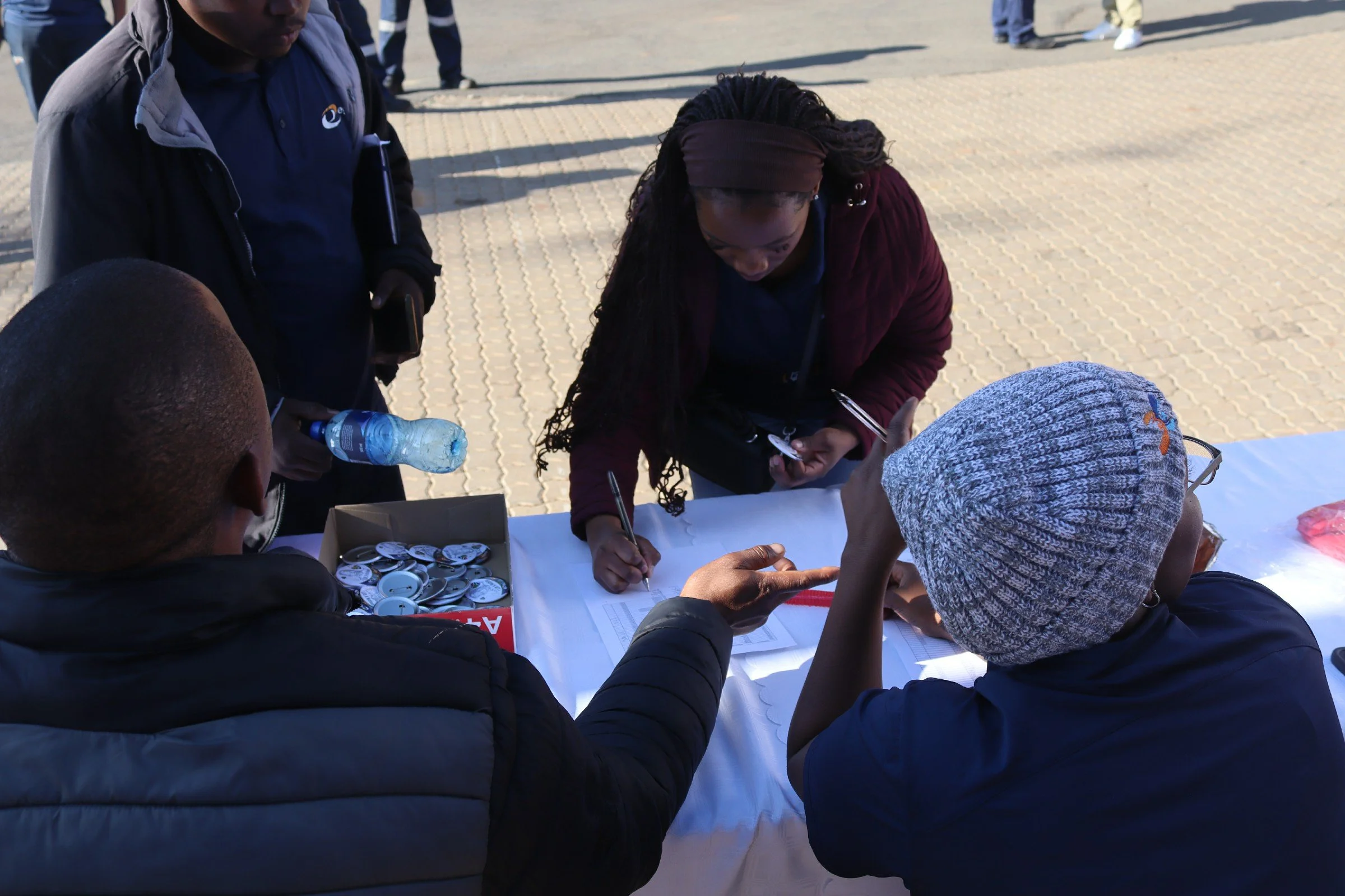 People gathered at a table outdoors, filling out forms, with buttons and a water bottle on the table.
