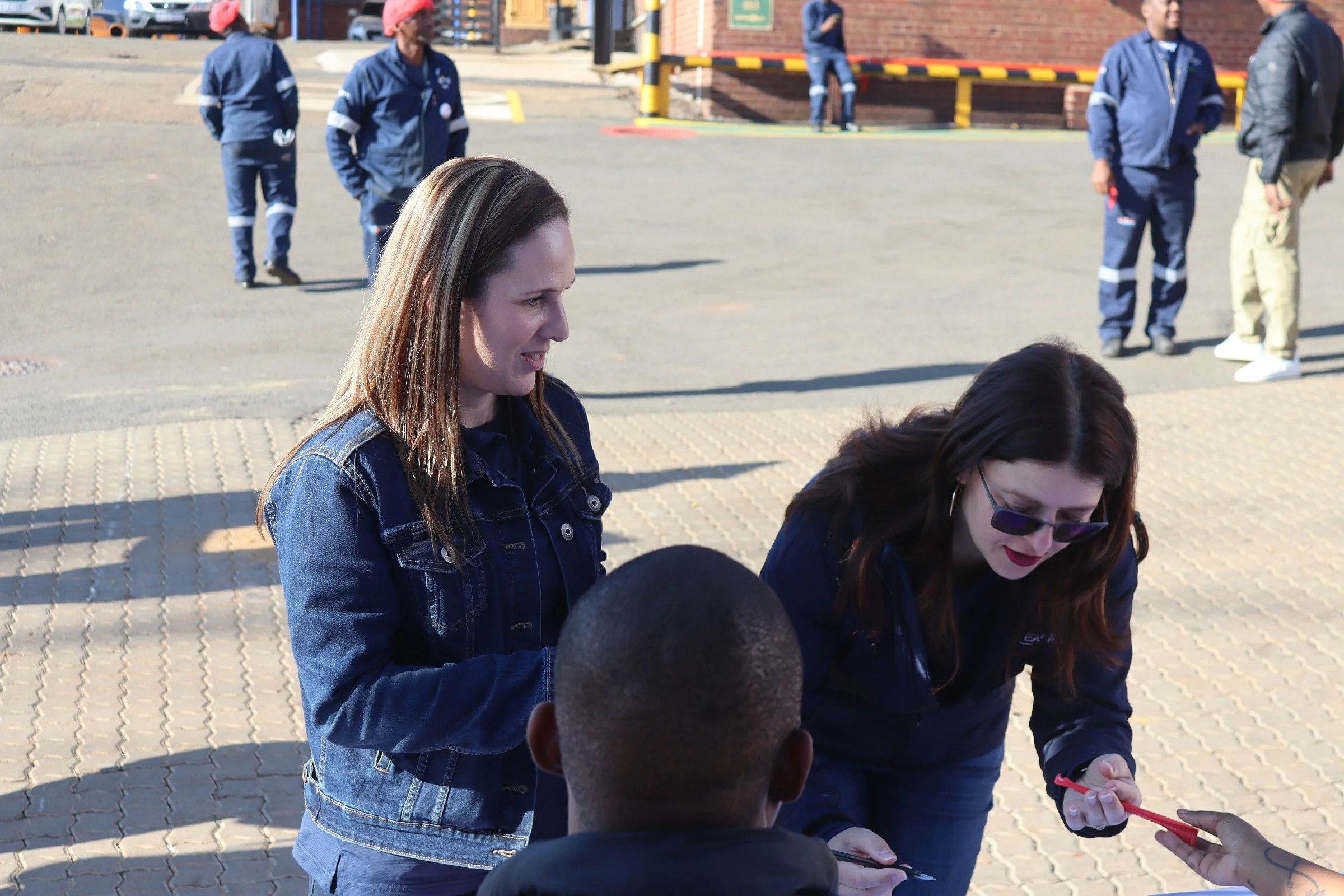 Group of women outdoors during daytime, engaging in interaction, with a brick building and several people standing in the background.