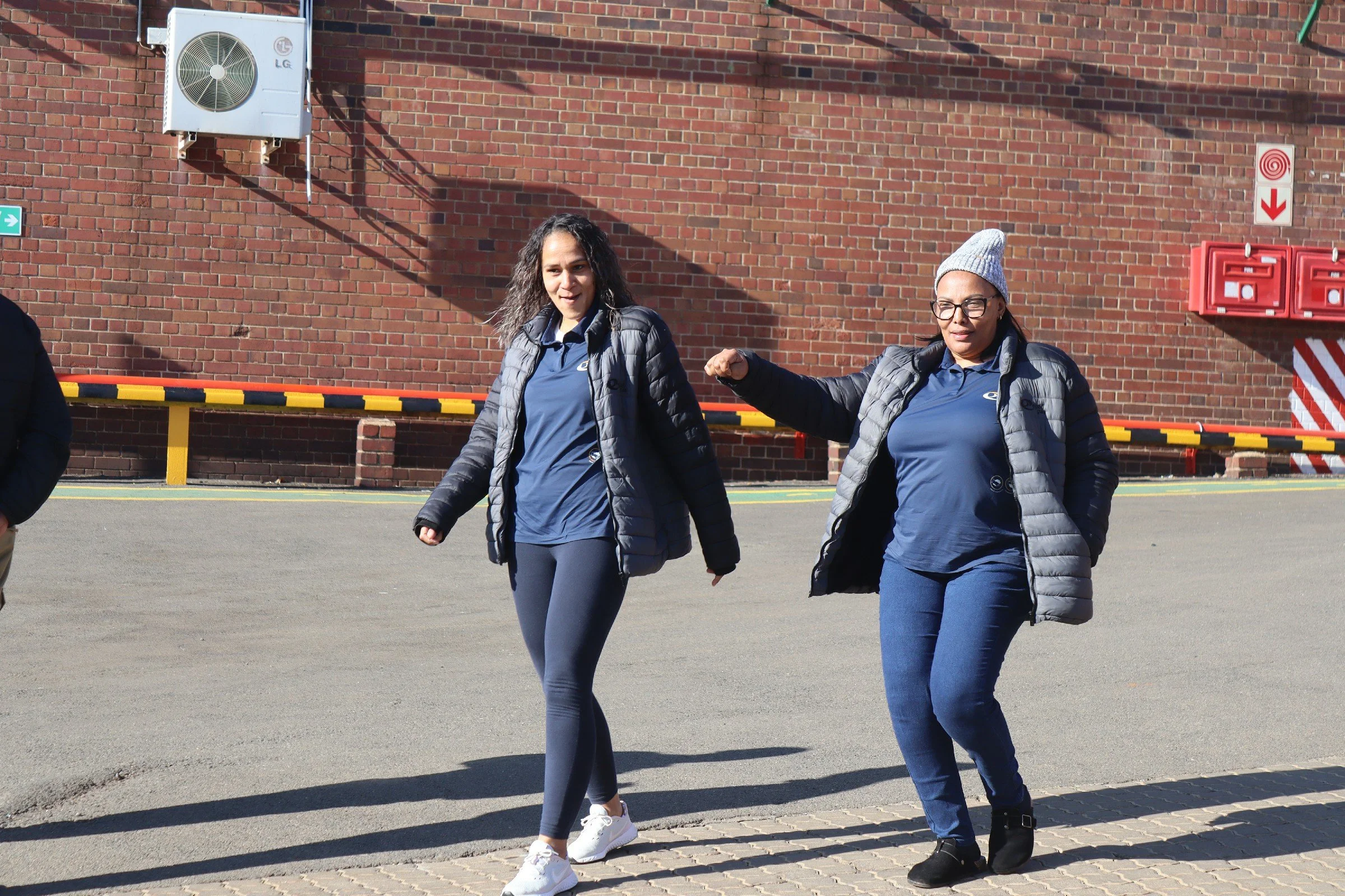 Two women walking outdoors near a brick building, one with curly dark hair and glasses, wearing a beanie and gray jacket, the other with straight dark hair, wearing a gray jacket and blue outfit.