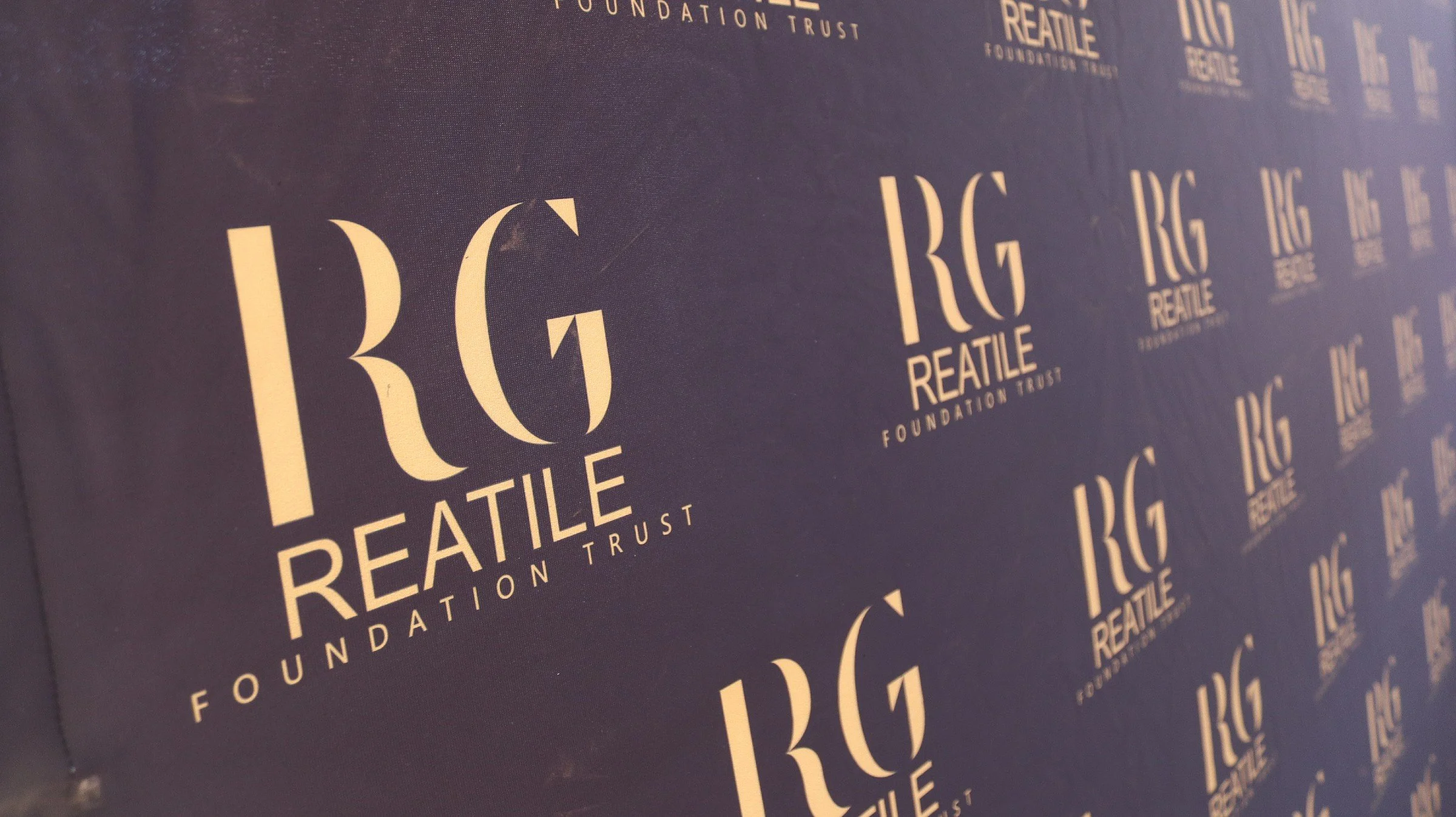 Close-up of a black fabric backdrop with repeated white text reading 'RG THE REATLE FOUNDATION TRUST' in a stylized font.