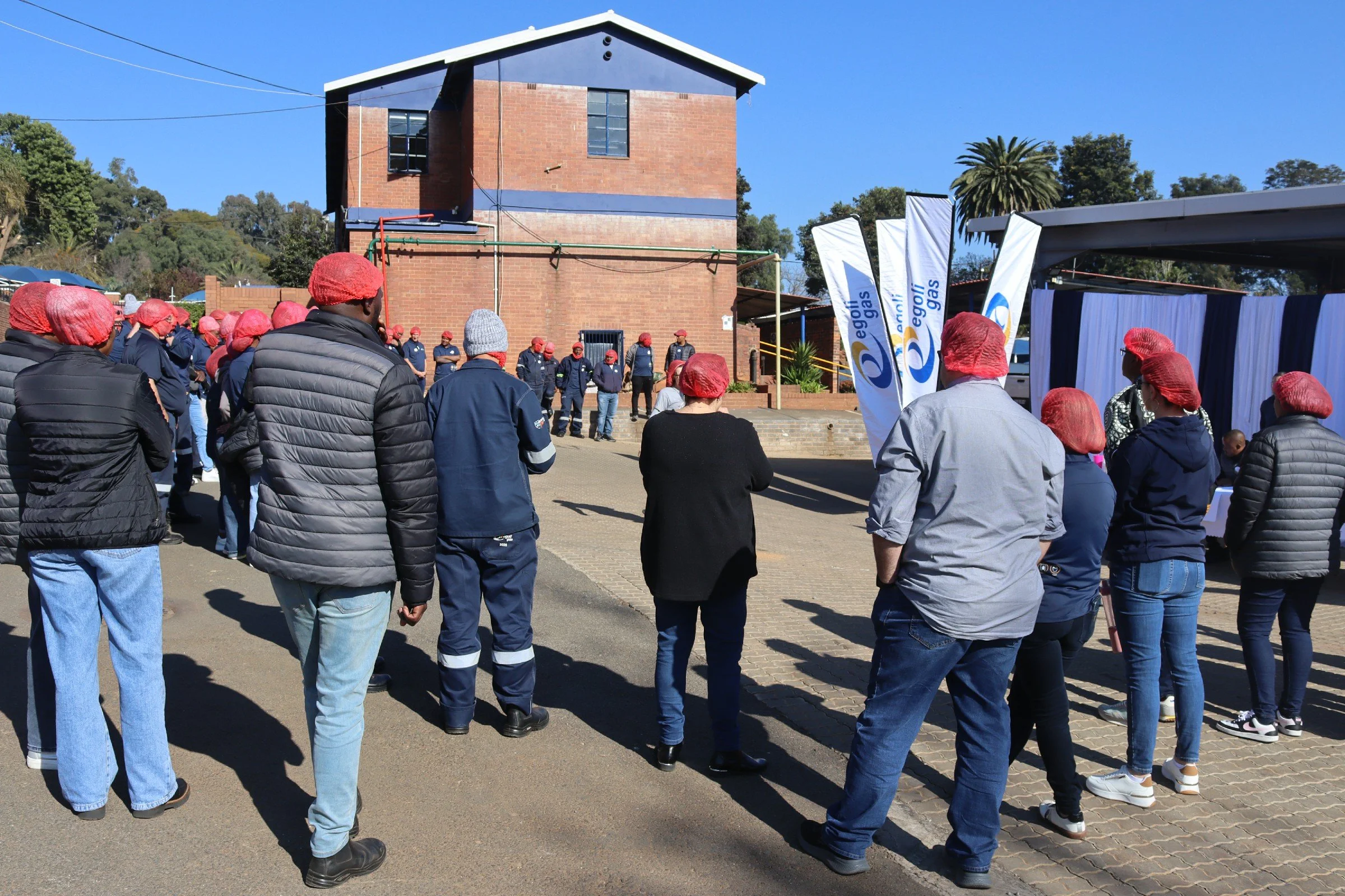 A gathering of people wearing red hairnets and dark jackets outdoors, standing in front of a brick building with flags and banners for Eskom Gas.