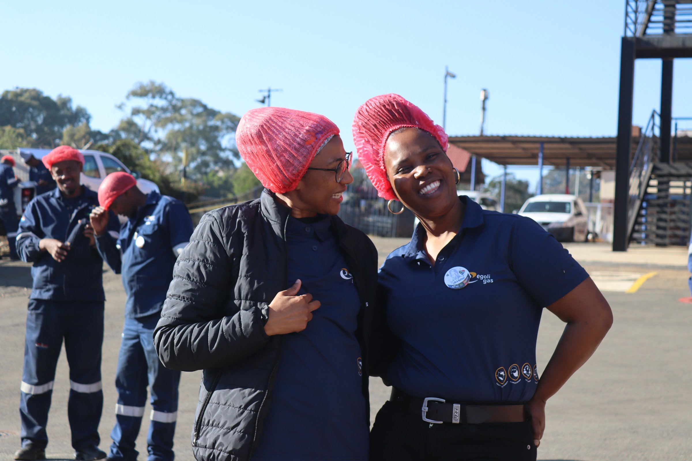 Two women wearing pink hairnets and dark blue shirts are smiling and talking to each other outdoors, with several other people in similar uniforms and red hairnets in the background.