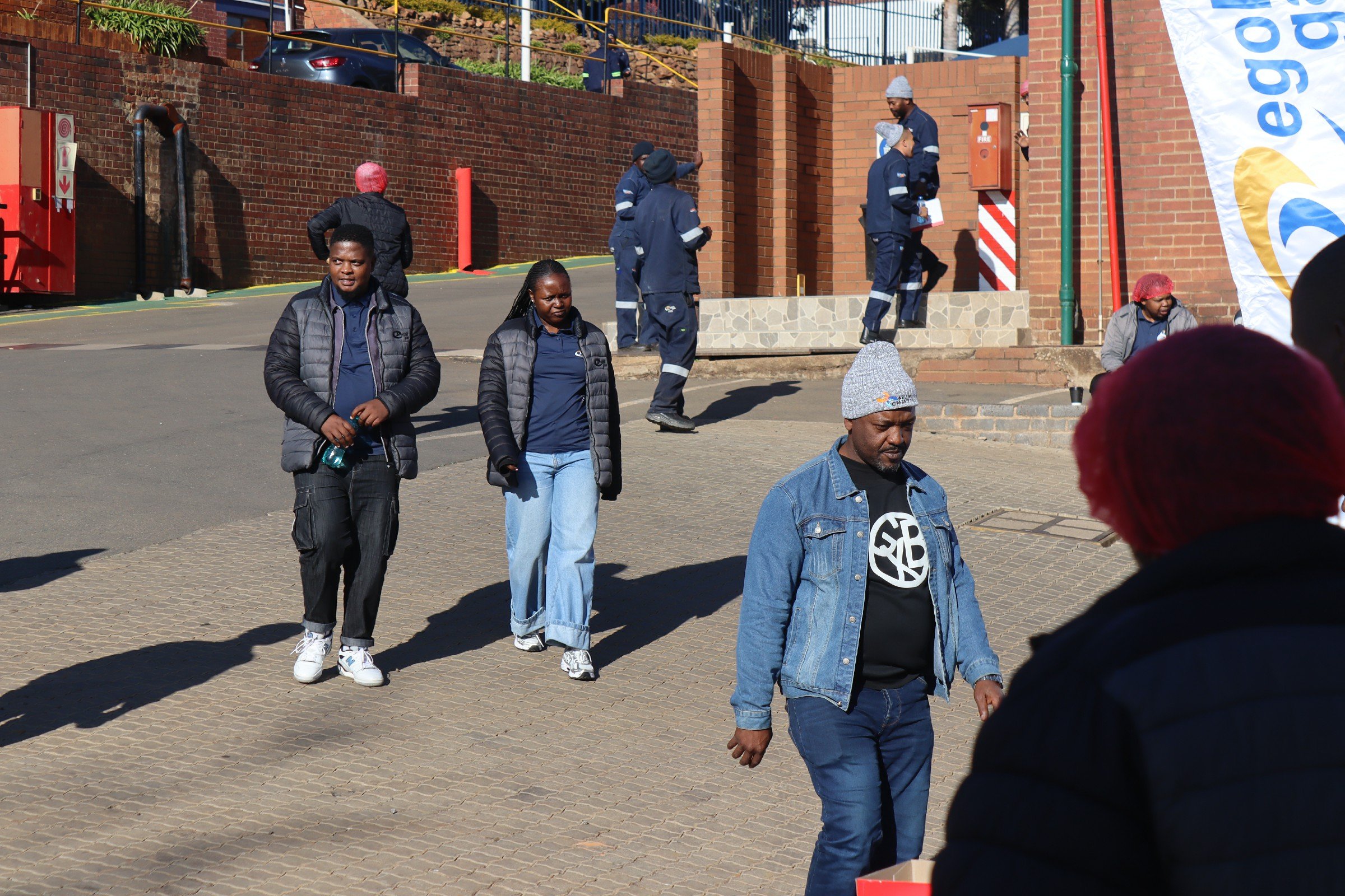 A group of people walking outside near a brick building with a small set of stairs. Some individuals are wearing black jackets, and one person is wearing a grey jacket. In the background, several people are standing on the stairs and appear to be eng