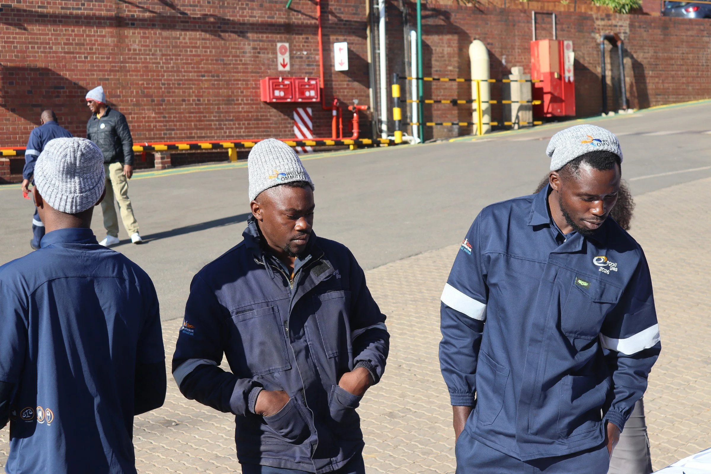 Group of workers standing outside near a brick wall, wearing navy blue uniforms and gray knit hats.