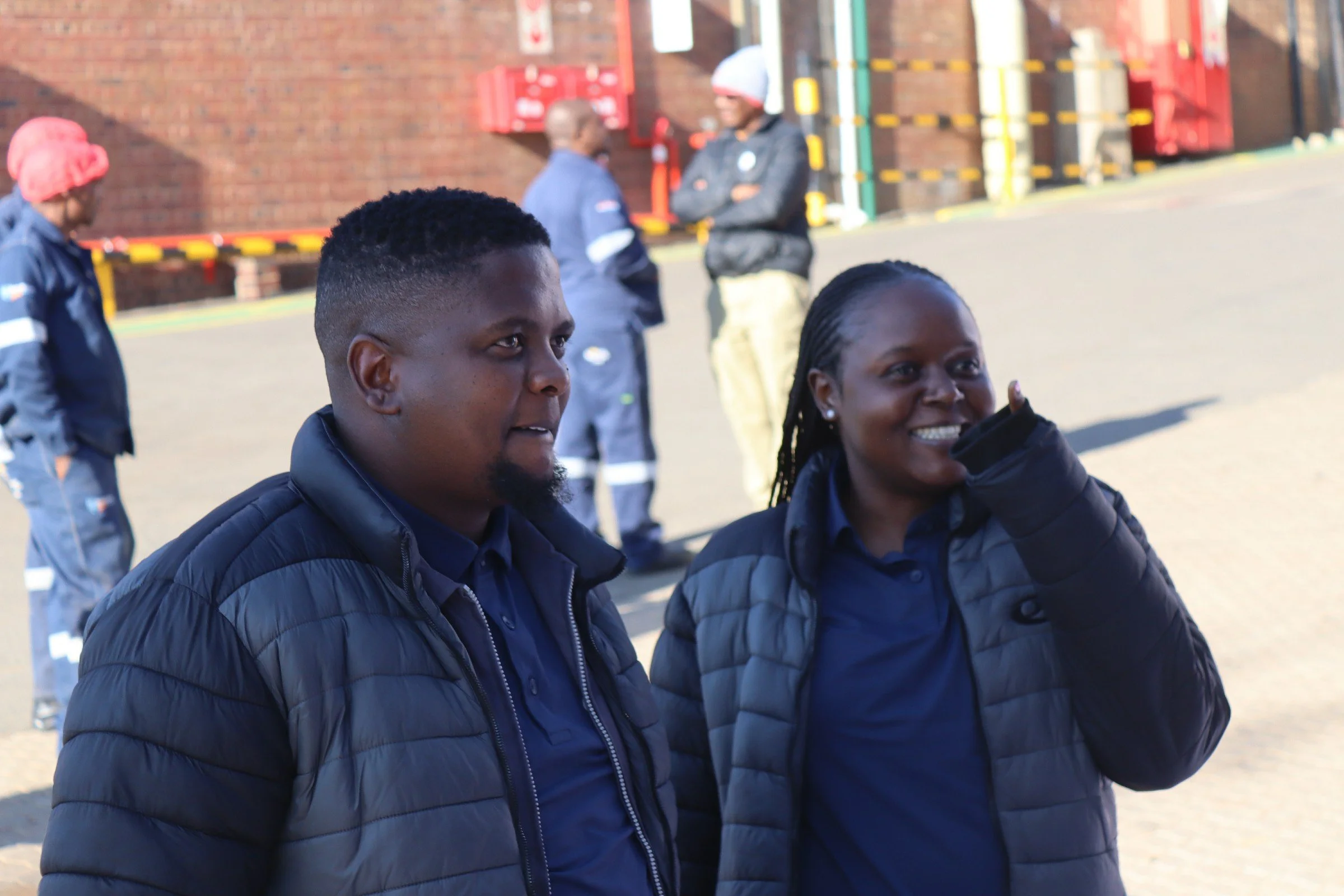 Two African American women smiling, wearing black jackets, standing outdoors with a brick building and other people in the background.