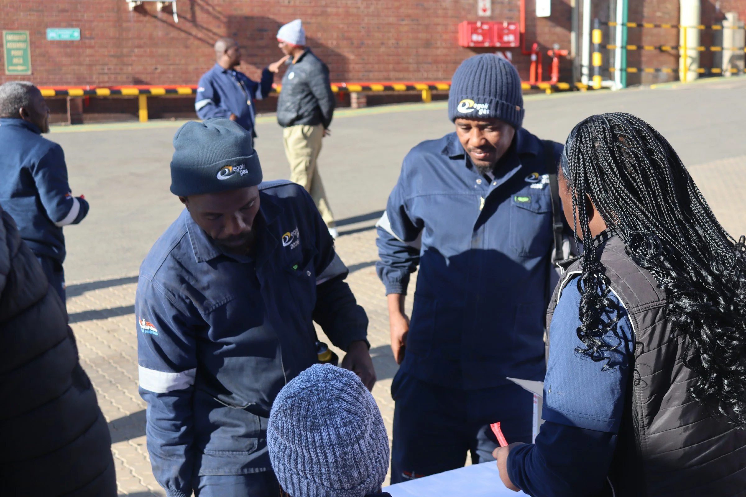 A group of people wearing blue uniforms and hats gathered outdoors, with some individuals engaged in conversation and one person pointing, near a brick building.
