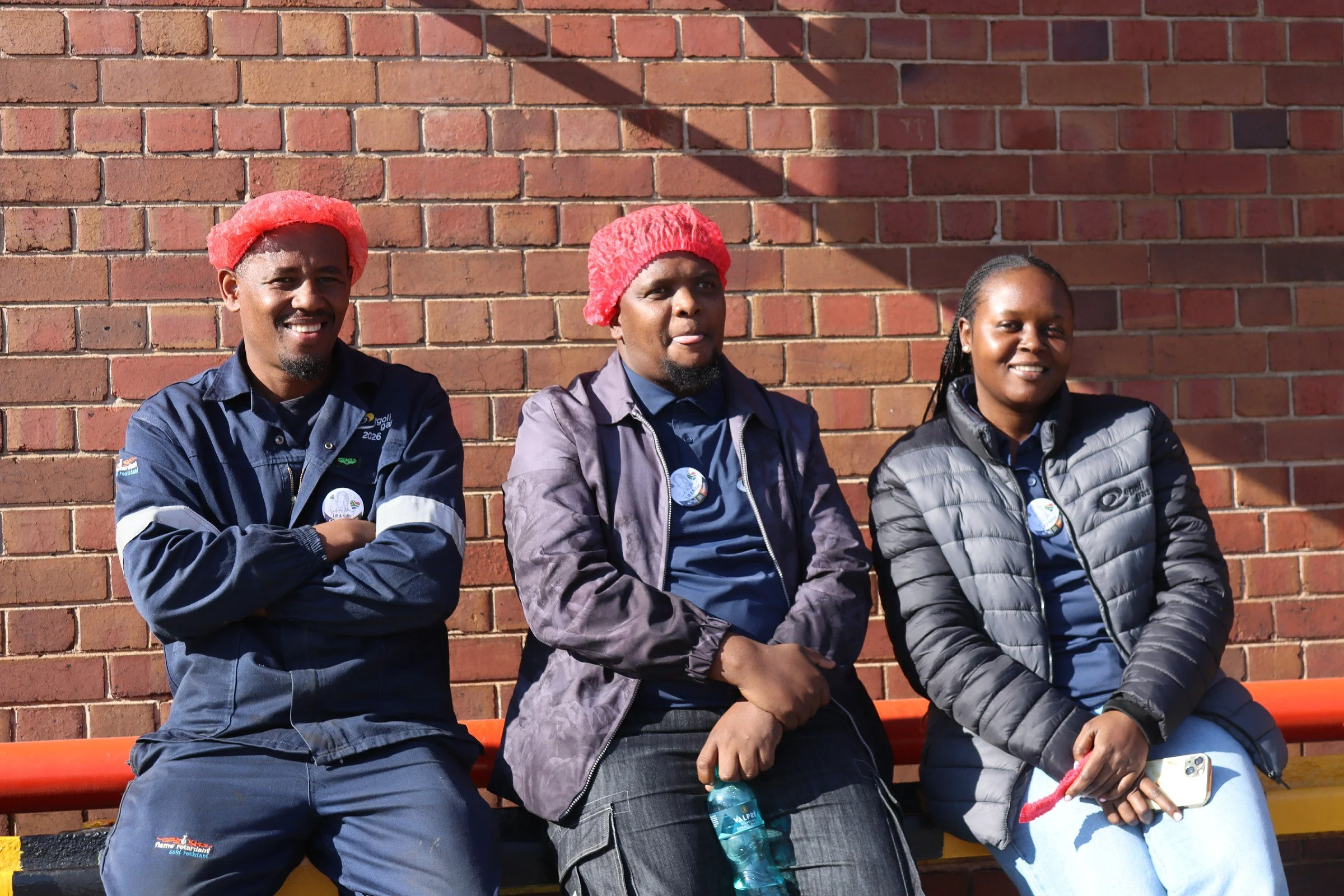 Three people sitting on a bench against a brick wall, wearing warm clothing and red hats, smiling and relaxed.