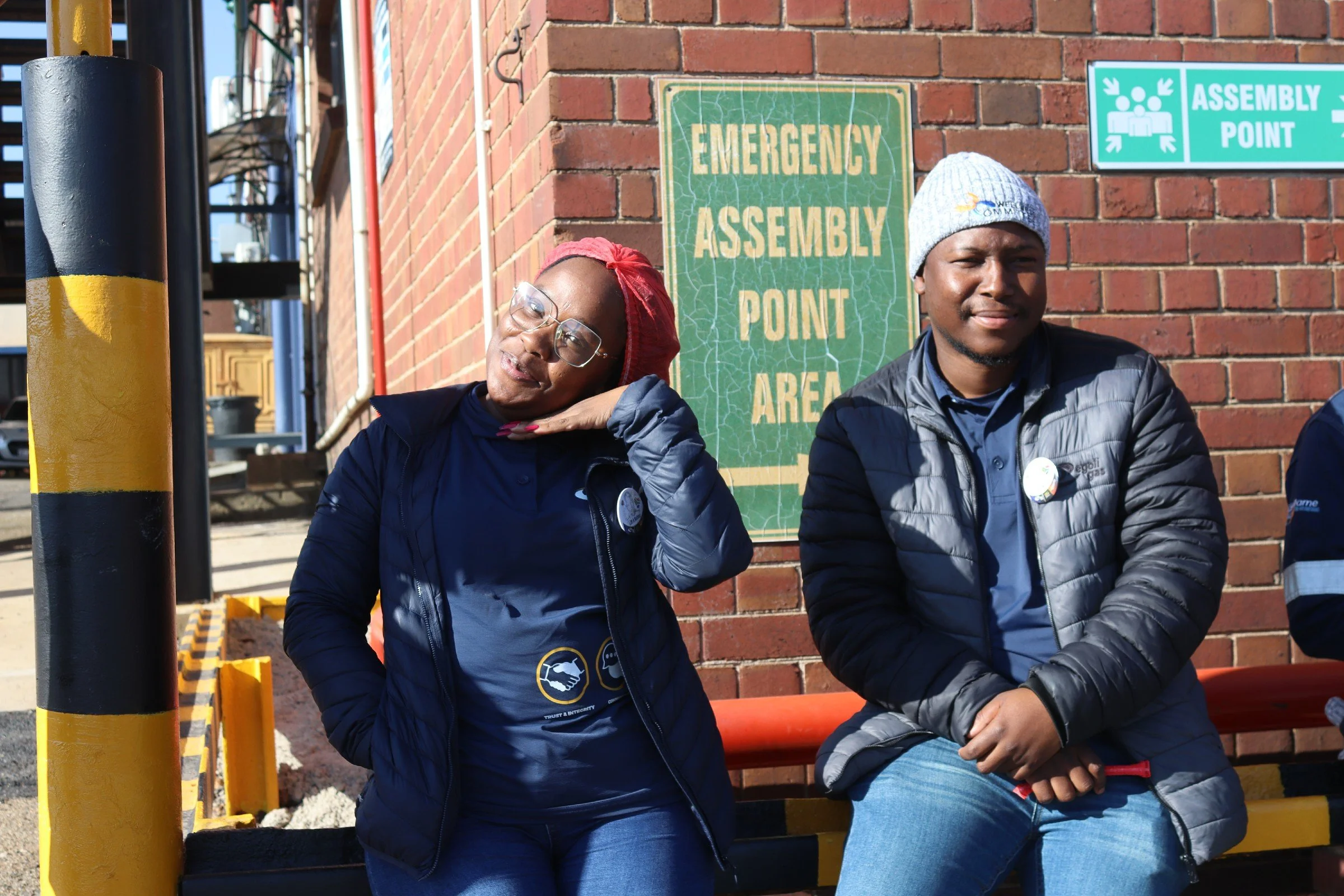 Two people sitting in front of a brick wall with signs that read 'Emergency Assembly Point' and 'Assembly Point'. The person on the left, wearing glasses and a red headscarf, is resting her head on her hand. The person on the right, wearing a gray be