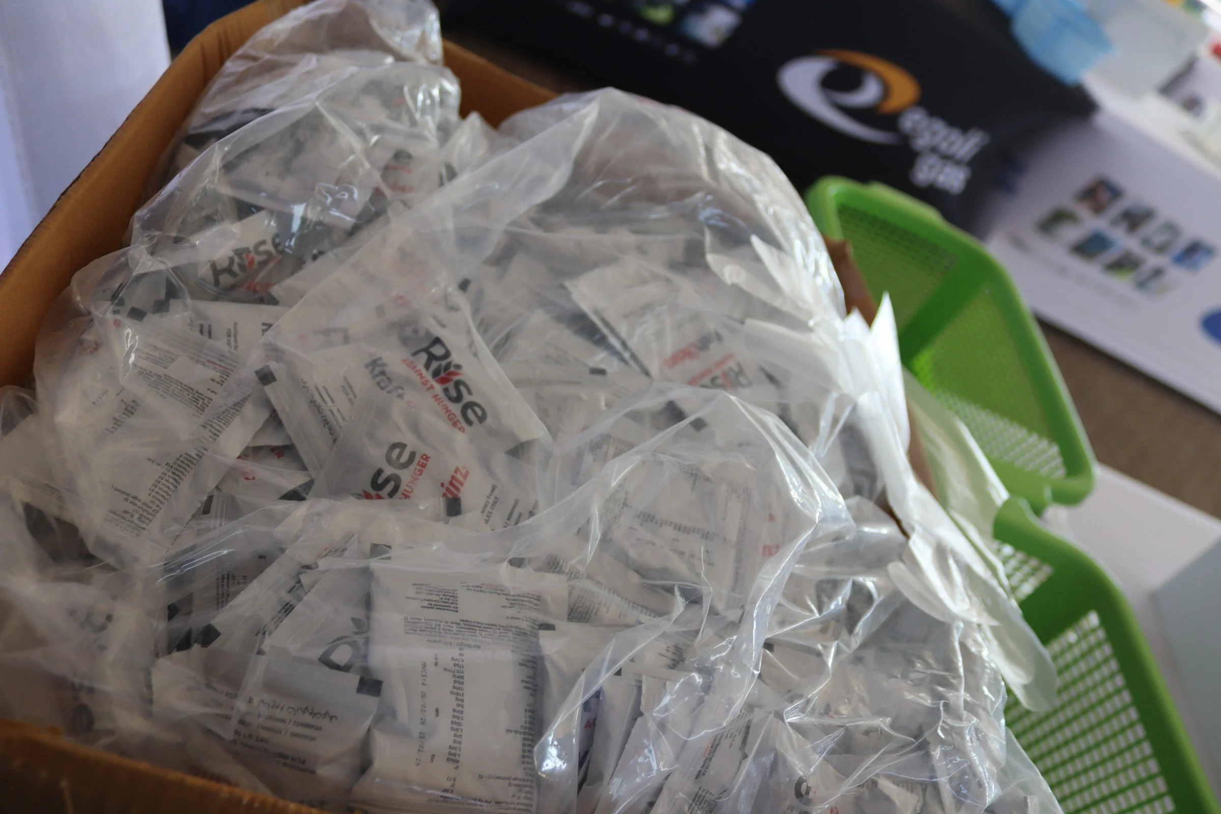 A cardboard box filled with clear plastic bags containing small items. In the background, part of a black box with a logo and some text, and a green plastic bin are visible.