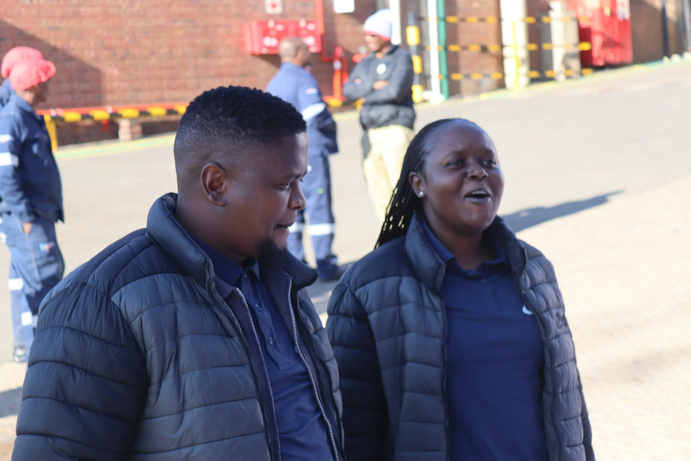 Two women wearing dark jackets standing outdoors with other people in the background, near a brick wall with equipment on it.