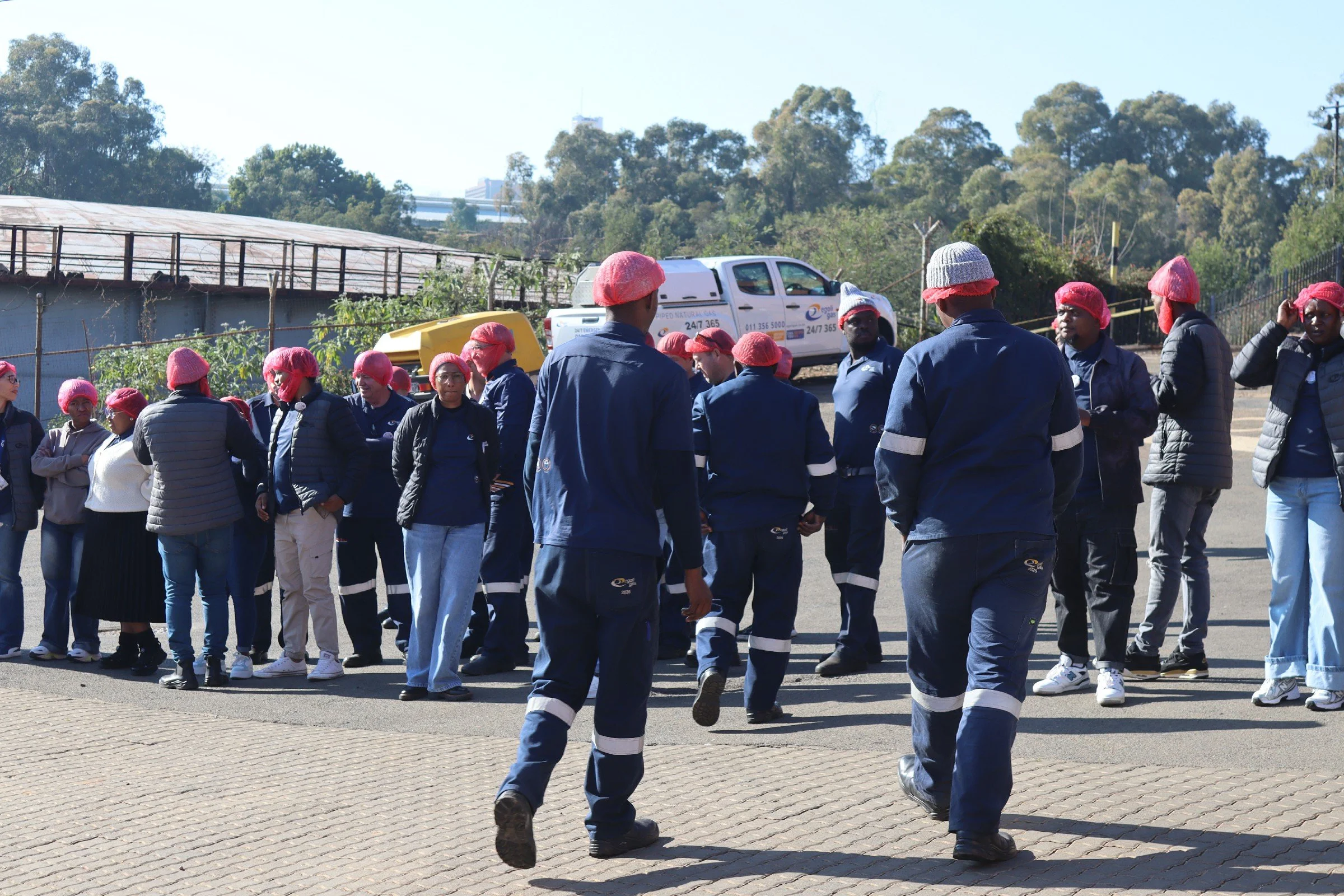 A group of people wearing matching dark blue uniforms and pink hairnets are standing outdoors on a paved area, possibly preparing for a team activity or event. They are engaged in conversation or listening to instructions, with some looking towards t