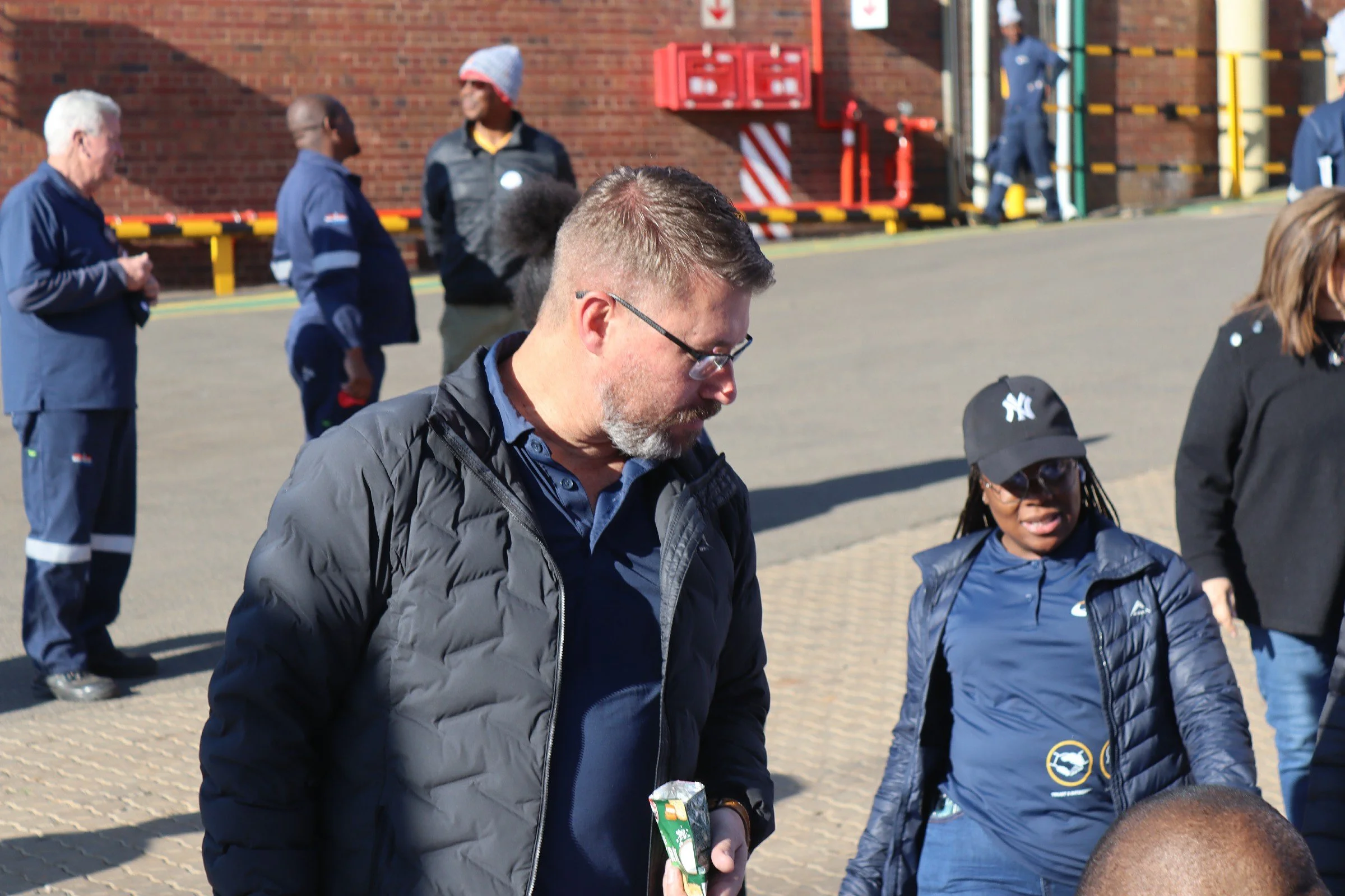 Group of people standing outdoors, some wearing dark blue jackets with white stripes, others in casual clothing, near a brick building with fire alarm boxes and safety barriers in the background.