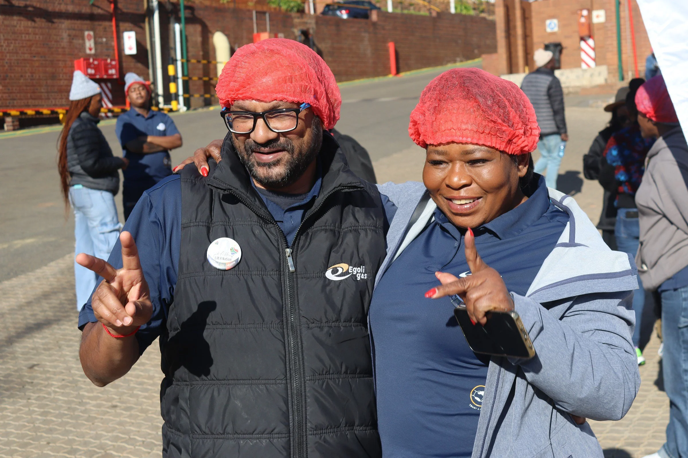 Two people with red hair nets smiling and making peace signs, outdoors in front of a brick wall with other people in the background.