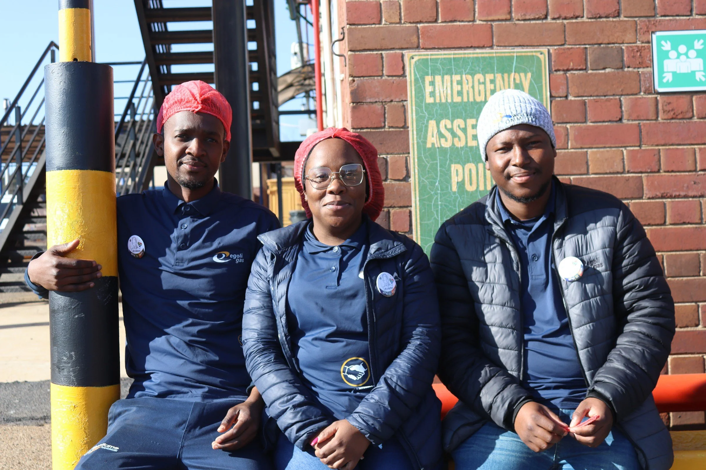 Three individuals sitting outdoors next to a yellow and black pole in front of a brick wall with an 'Emergency Assembly Point' sign. They are dressed in dark jackets and red or gray head coverings, and are looking at the camera.