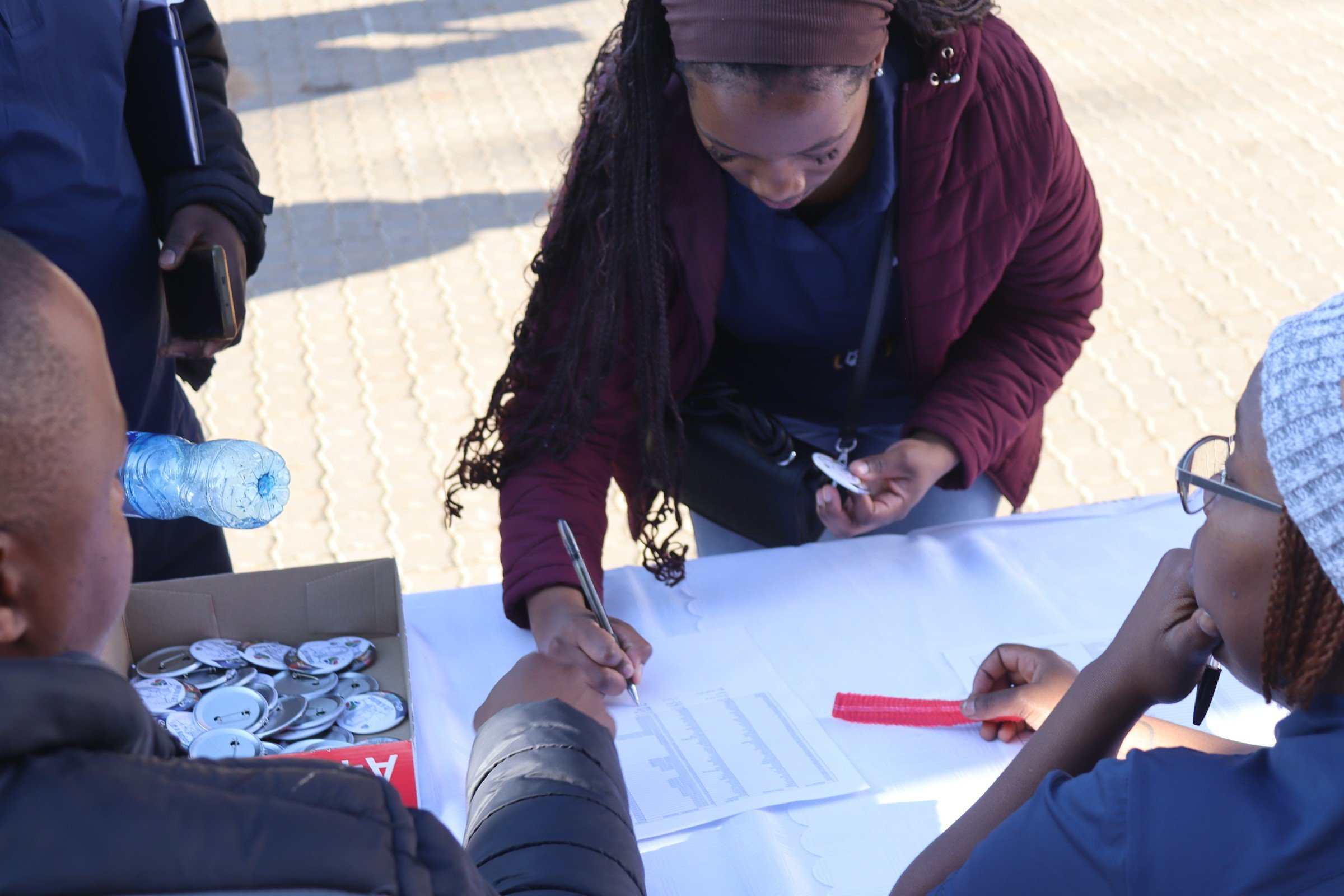 A group of people gathered around a table outdoors, signing papers, with badges and badges with buttons on the table, and a clear plastic water bottle, in a sunny setting with cobblestone ground.