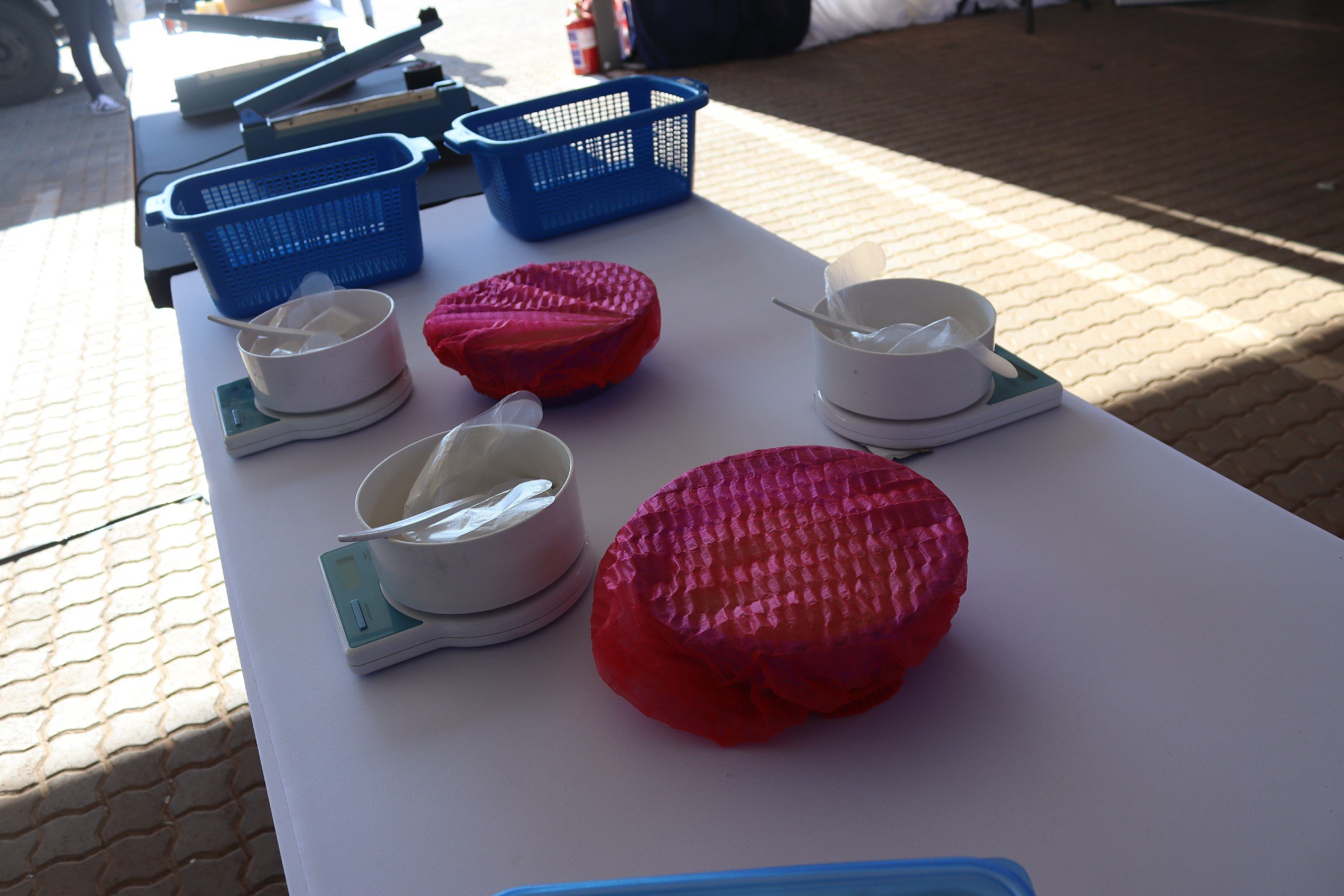 Laboratory setup with digital scales, pink mesh filters, and blue baskets on a white table, outdoors on brick pavement.