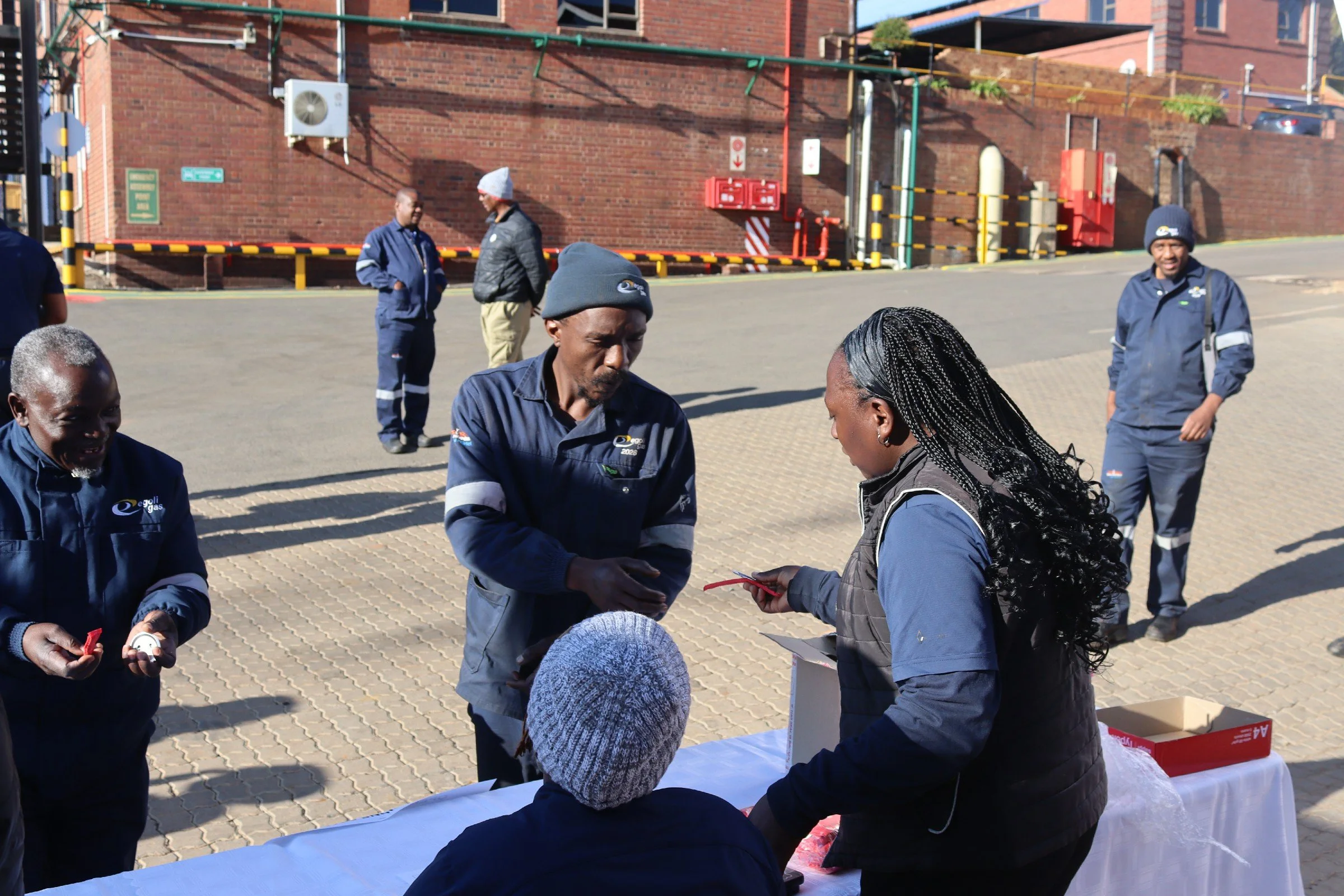 A group of people, including some in blue uniforms, gather outdoors near a brick building with a parking lot in the background. Some individuals are engaged in conversation, and there are tables with papers or items on them.