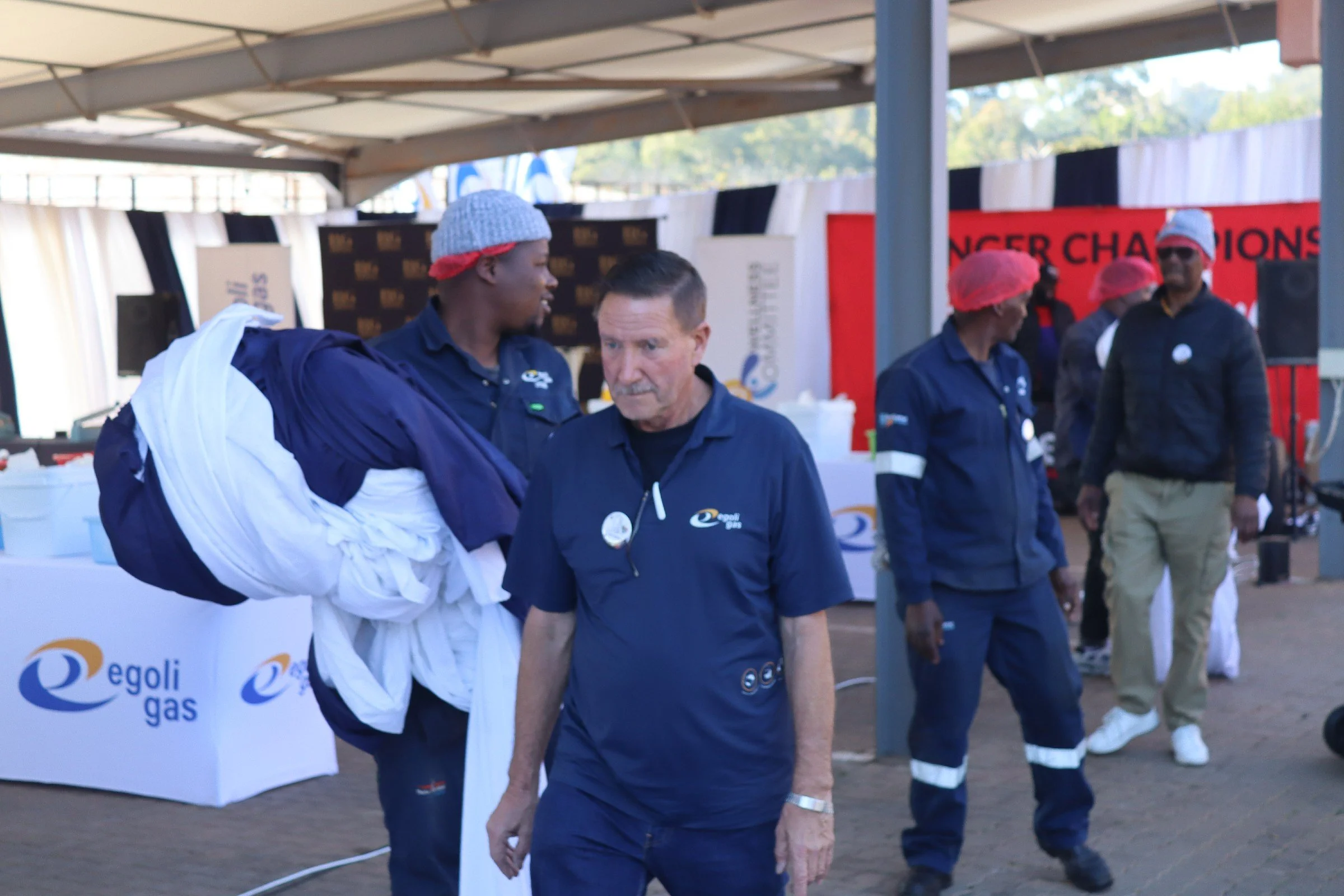 People wearing dark blue shirts and red hairnets are at an outdoor event under a canopy. One man is holding a white jacket, and others are engaged in conversation, with signage reading 'Egol gas' visible in the background.