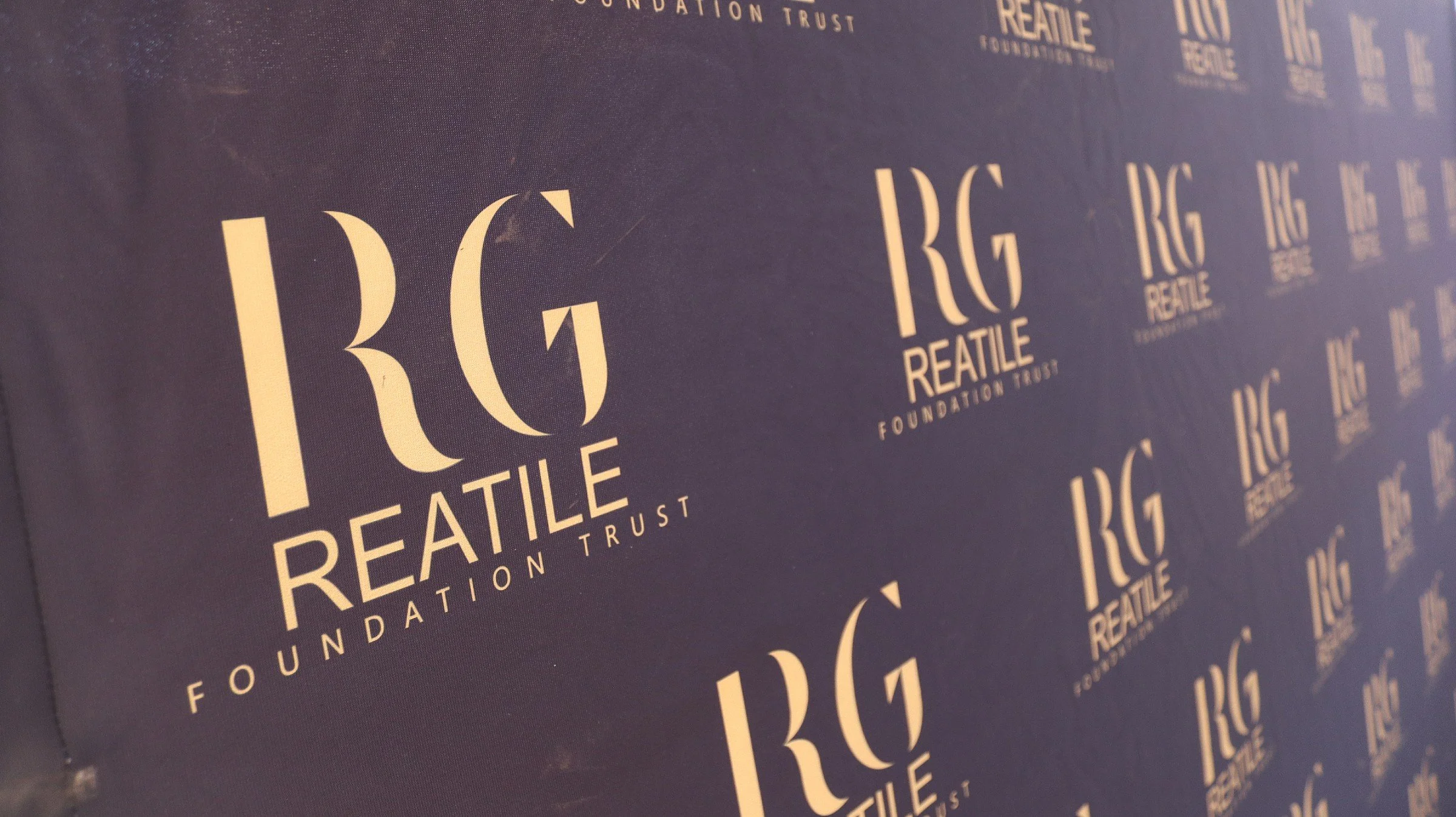 Close-up of a repeated logo for the REATILE Foundation Trust on purple fabric background.