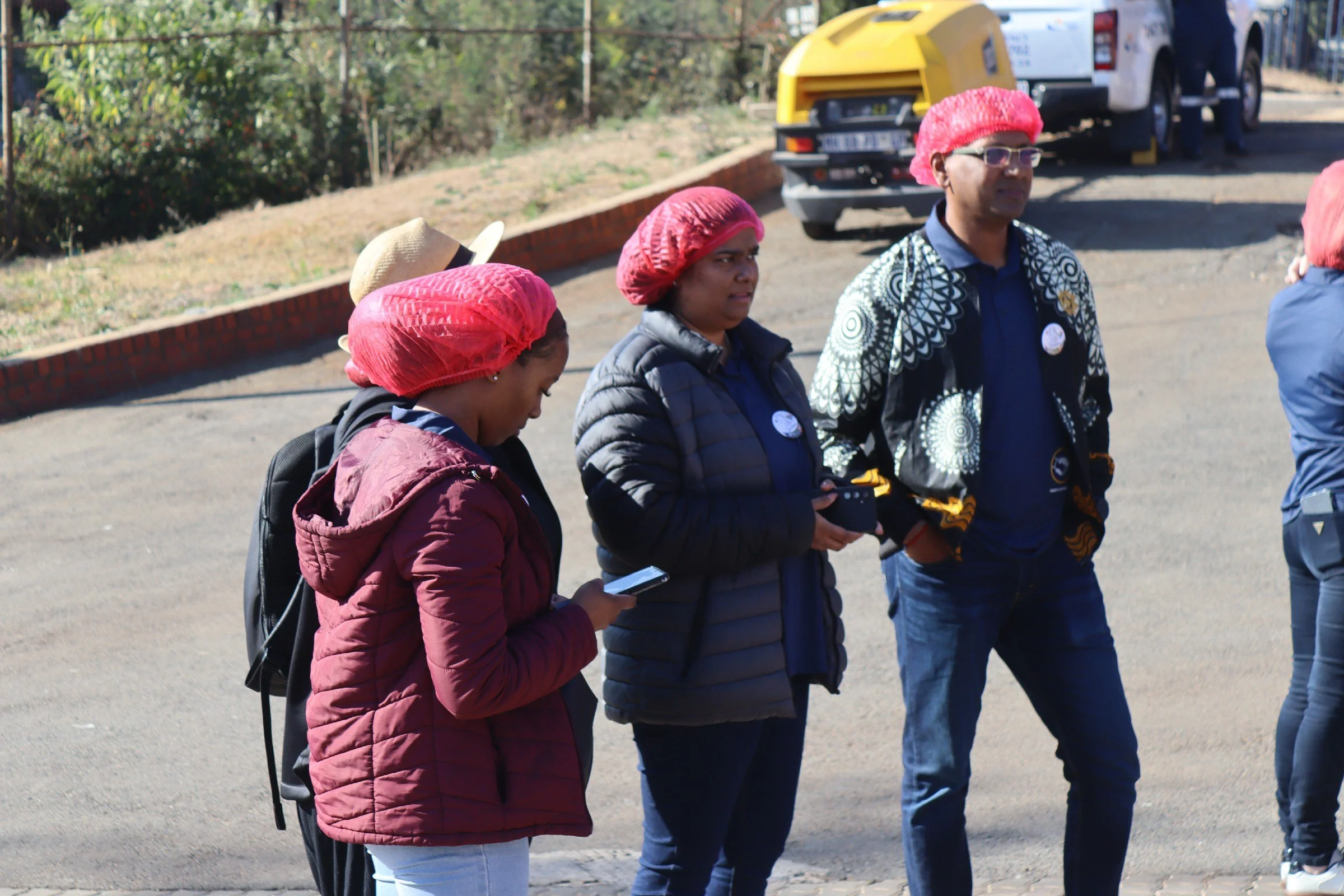 Group of people standing outdoors on a sidewalk. Some are wearing red hair nets, jackets, and holding phones, with a background of parked vehicles and greenery.