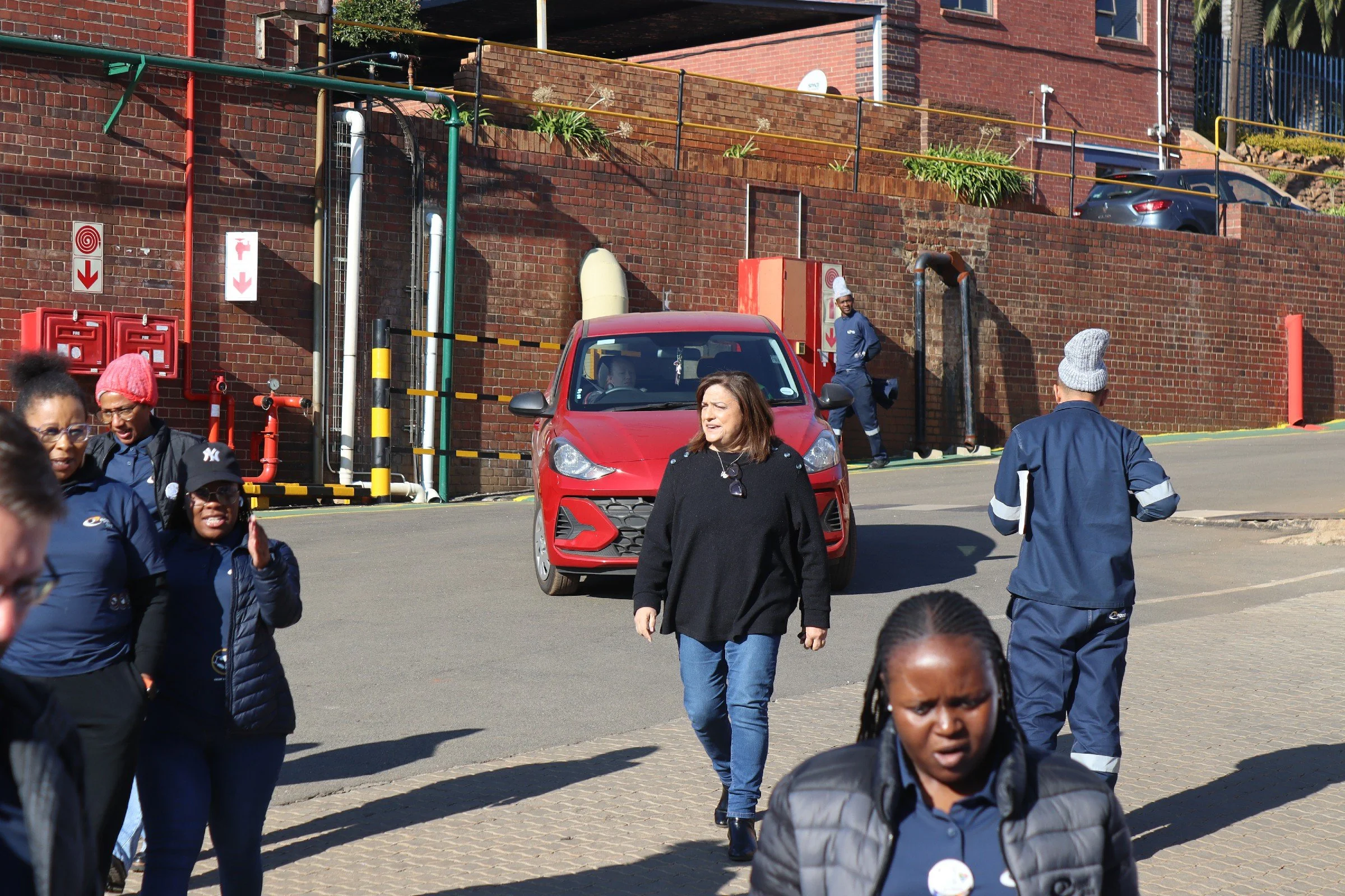 A group of diverse people walking outdoors near a red brick building with plants and parked cars.