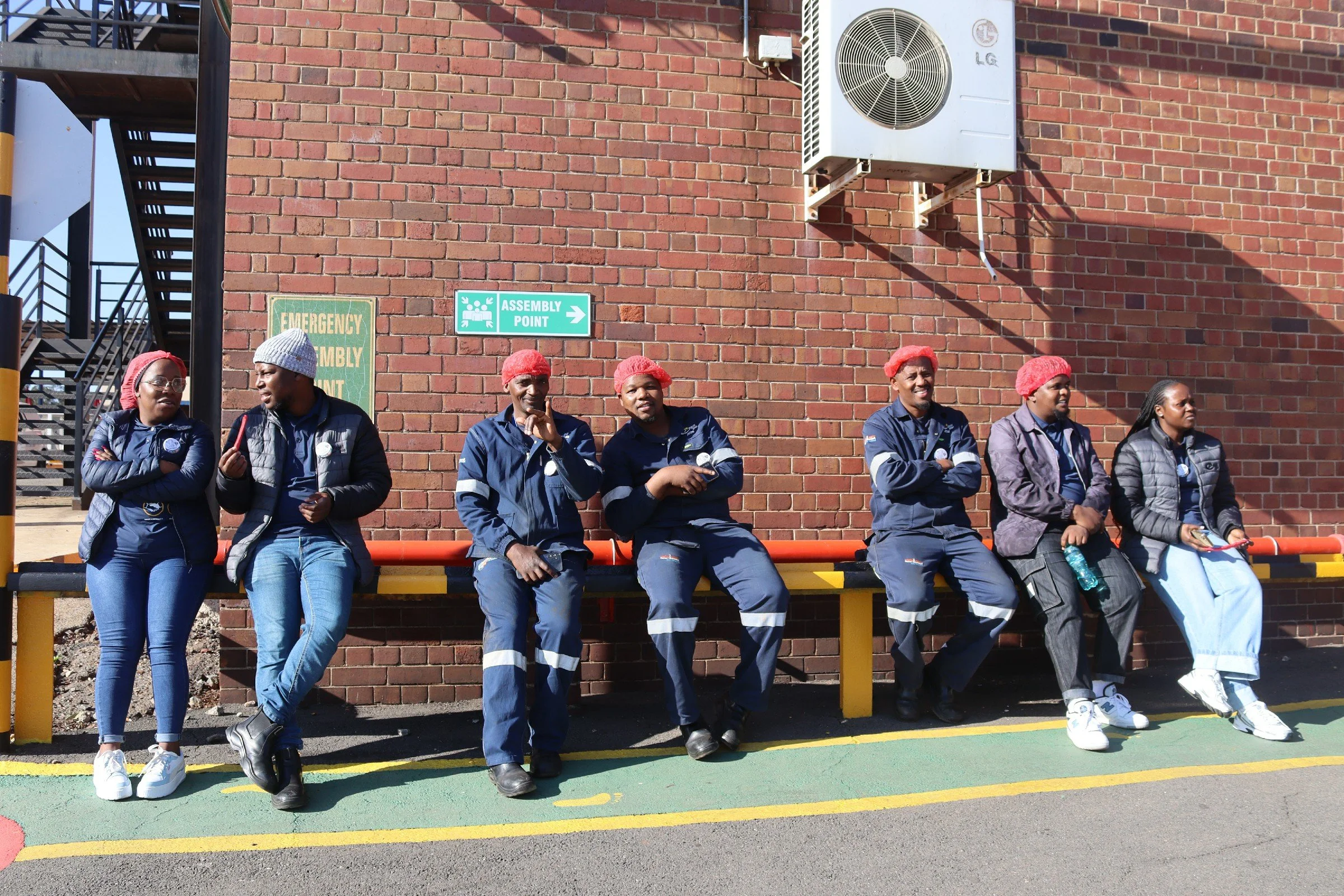 Seven people sitting on a bench outside a brick building, some with red hairnets, wearing blue work uniforms, and two women in casual clothing.