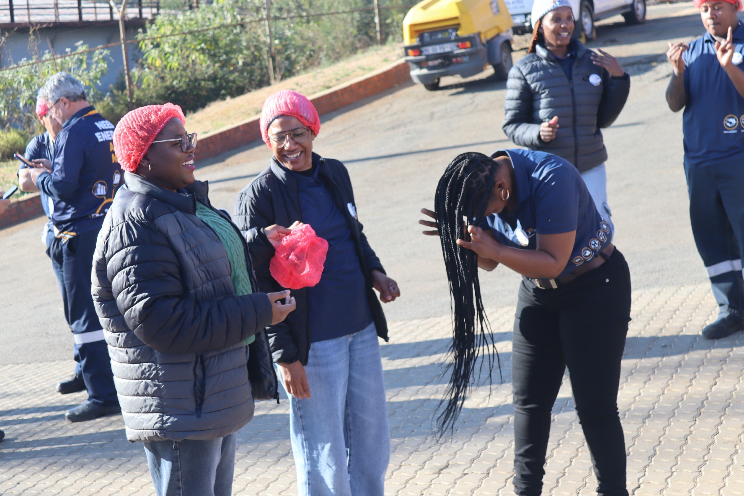 A group of smiling women wearing warm clothing and red or gray hair coverings stand and talk outdoors on a sunny day, one woman bending over and touching her hair.