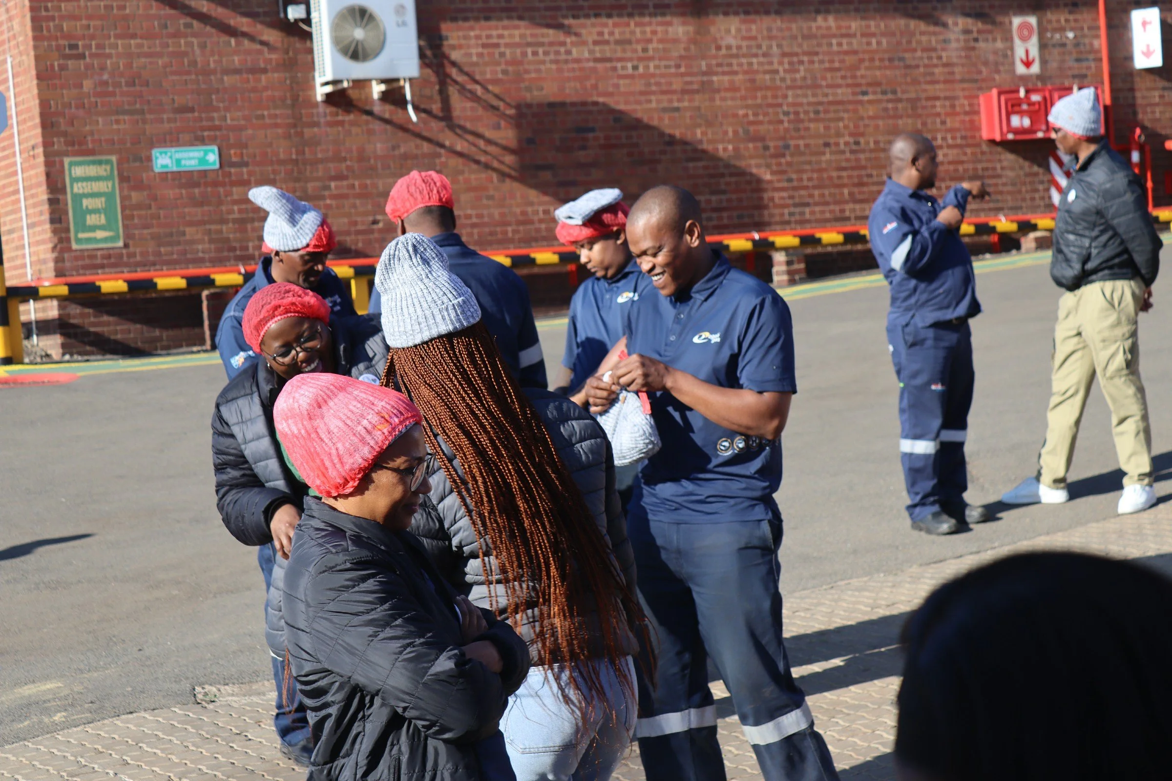 A group of people standing in a line outdoors near a brick building. Some are wearing red and gray knit hats, and everyone appears to be engaged or talking to each other.
