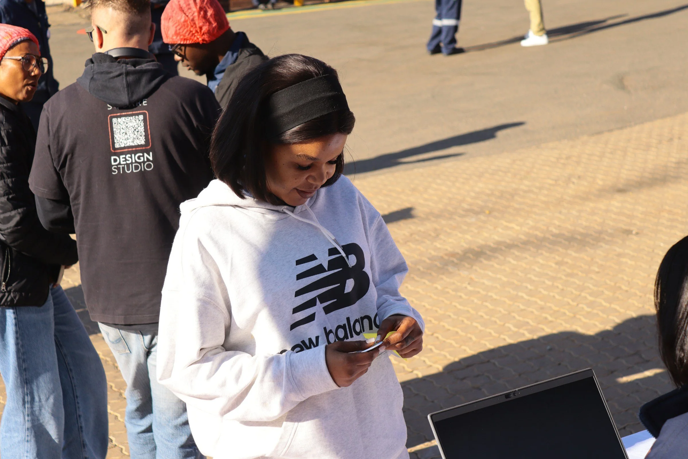 A woman with black hair wearing a black headband and a white hoodie with black text is looking at her phone. Several people are in the background, standing on an outdoor paved area.