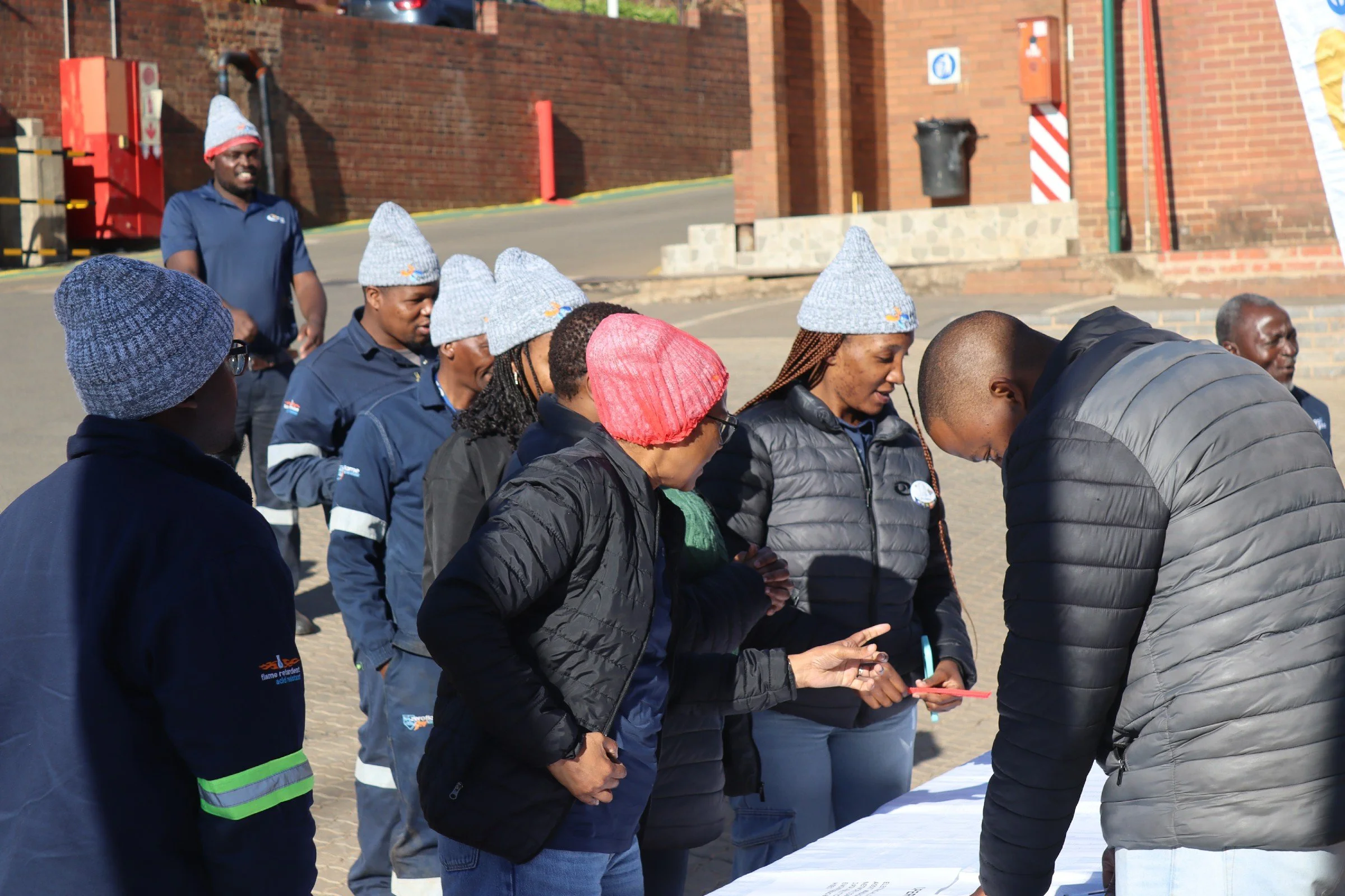 A group of people, diverse in gender and ethnicity, are gathered outdoors, bowing their heads in prayer or reflection. They are dressed in casual clothing and winter hats. The setting appears to be an urban area with brick buildings and street signs 