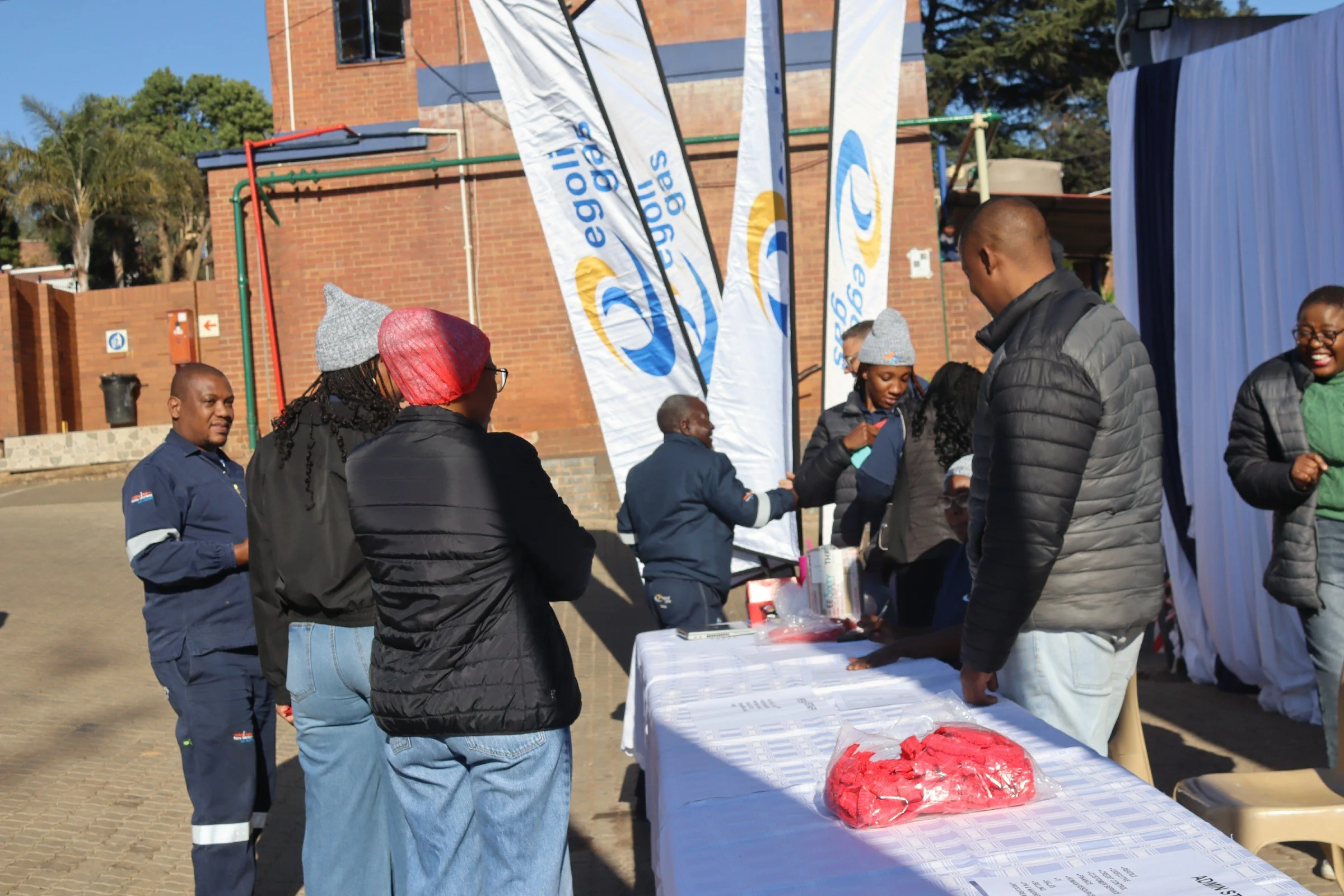 People gathered at an outdoor event, standing around a table with items on display, with flags in the background and a brick building behind them.