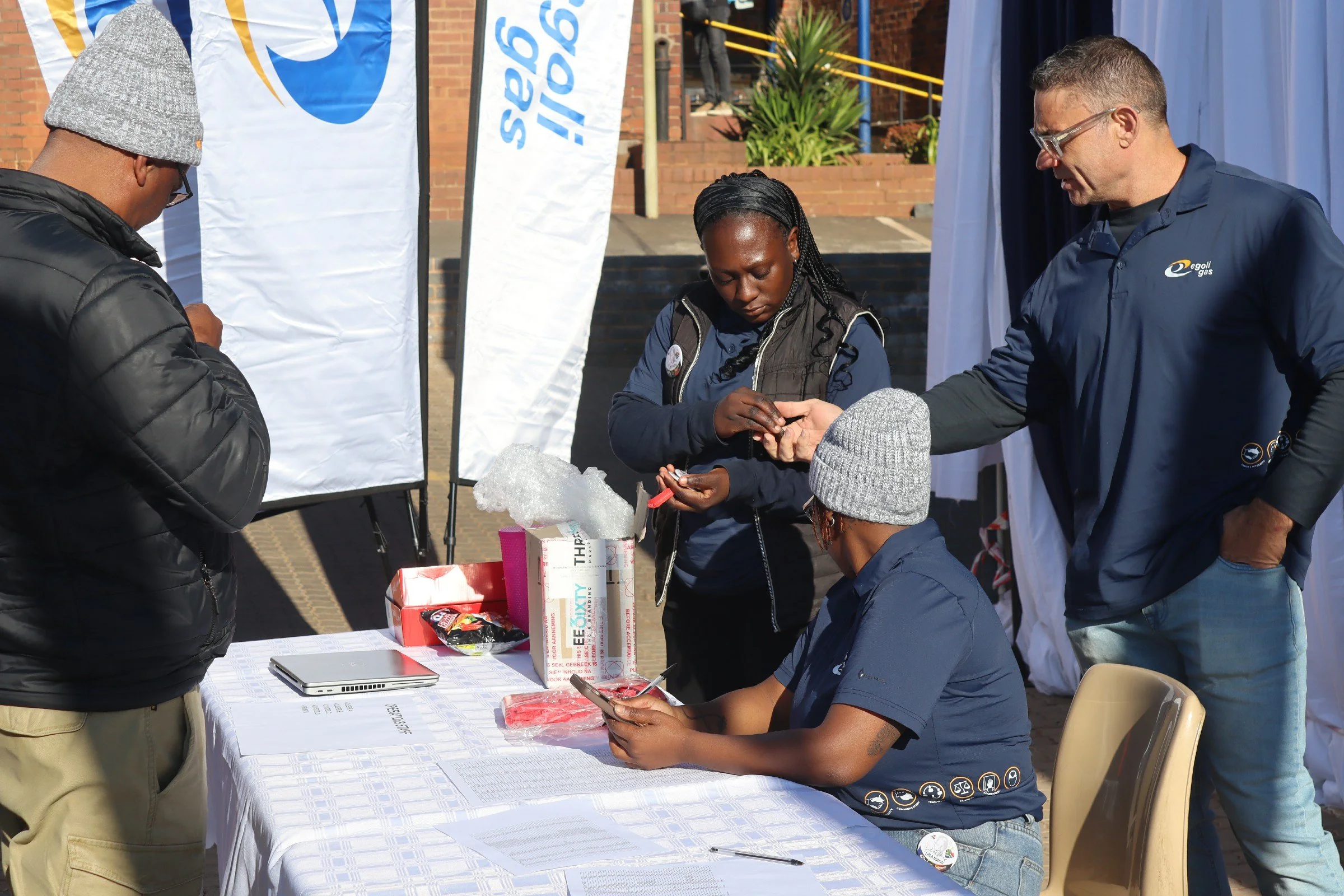 Group of people at an outdoor event around a table with informational materials, banners in the background, and one person holding a phone.