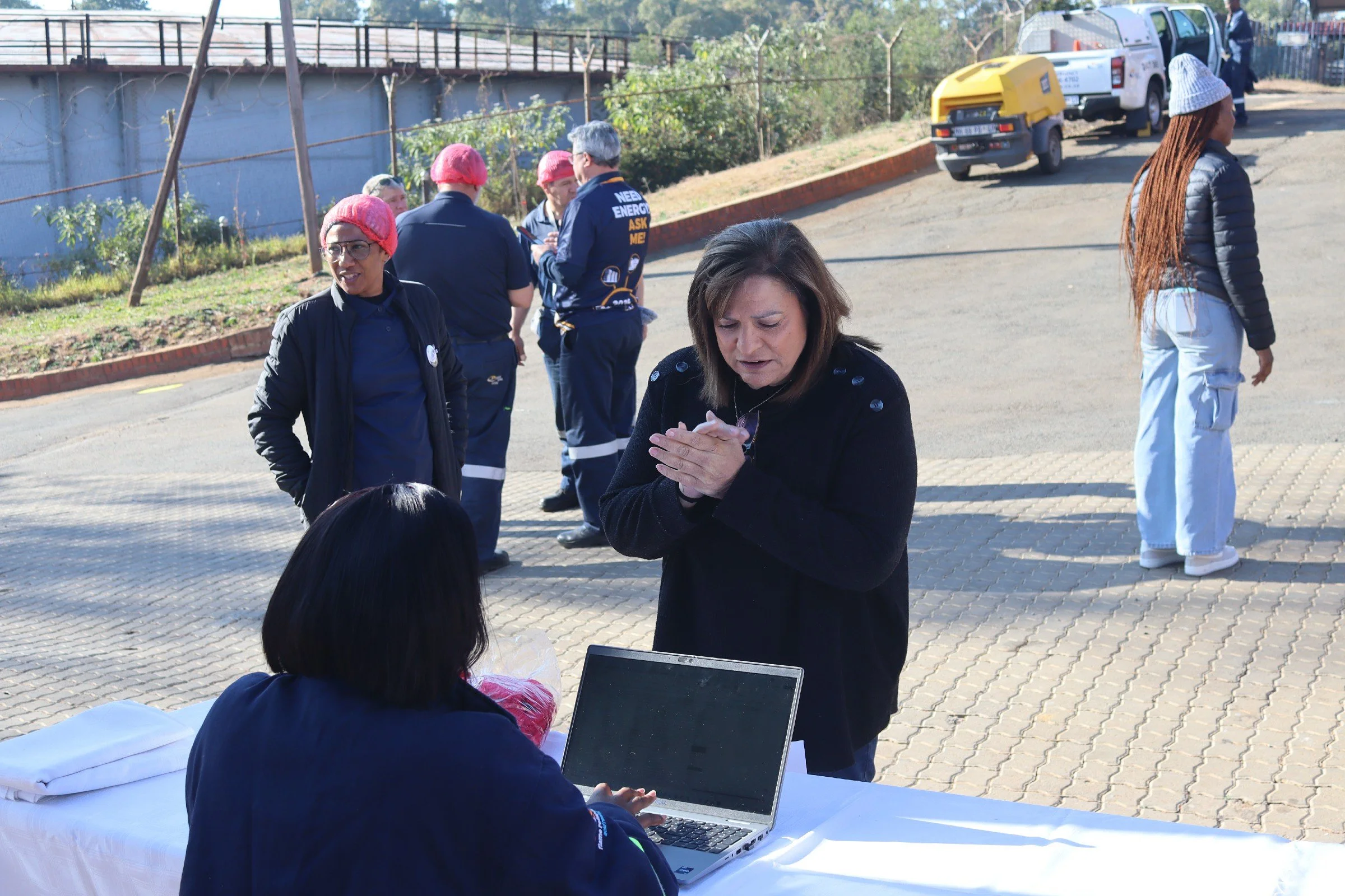 Group of people gathered outdoors near a chain-link fence, some wearing pink caps, engaged in conversation and working on a laptop at a table with a white tablecloth.