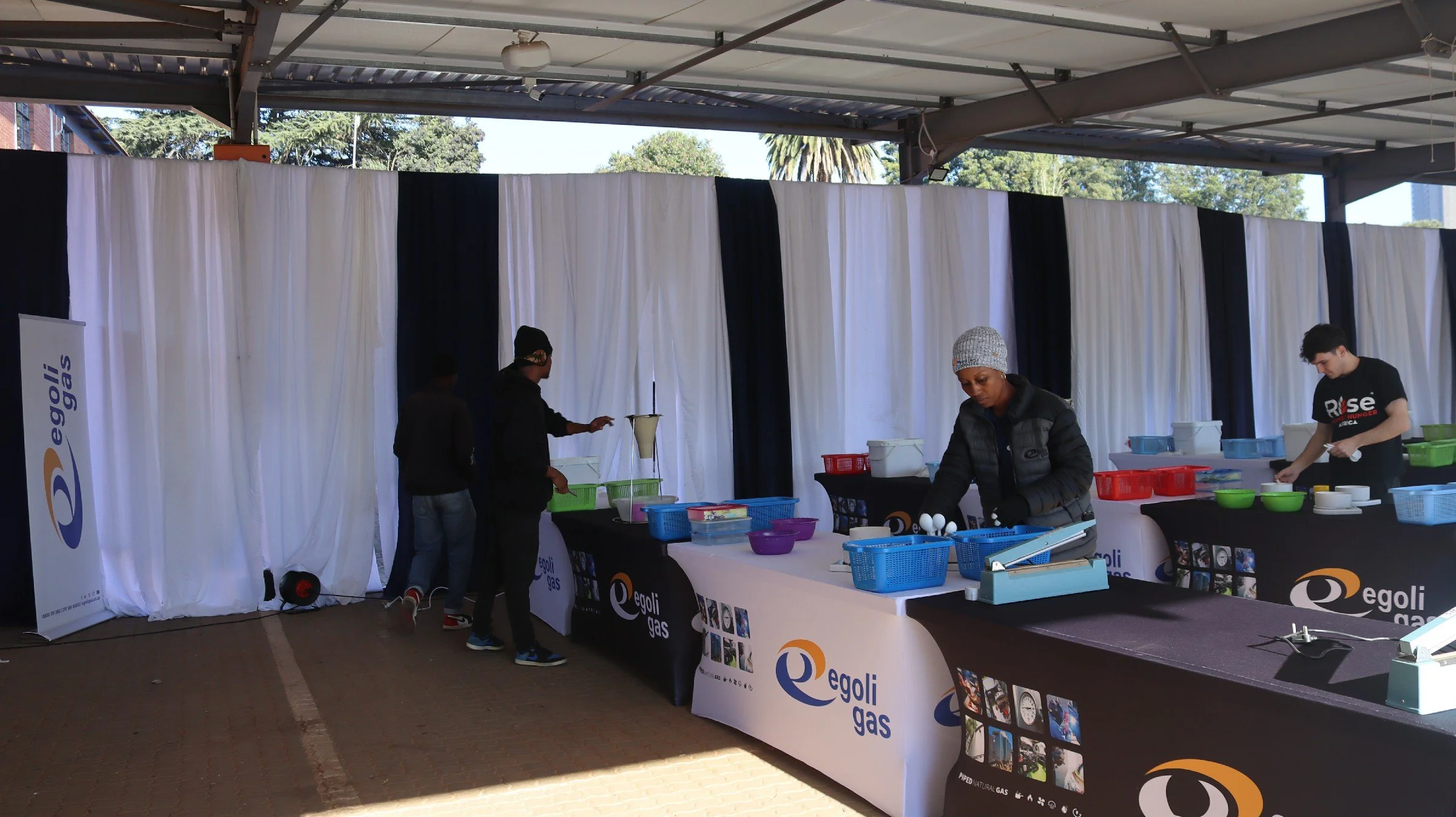 Indoor booth with white and black curtains, displaying Egoli Gas branding, with several people serving and preparing food at tables with bowls and utensils.