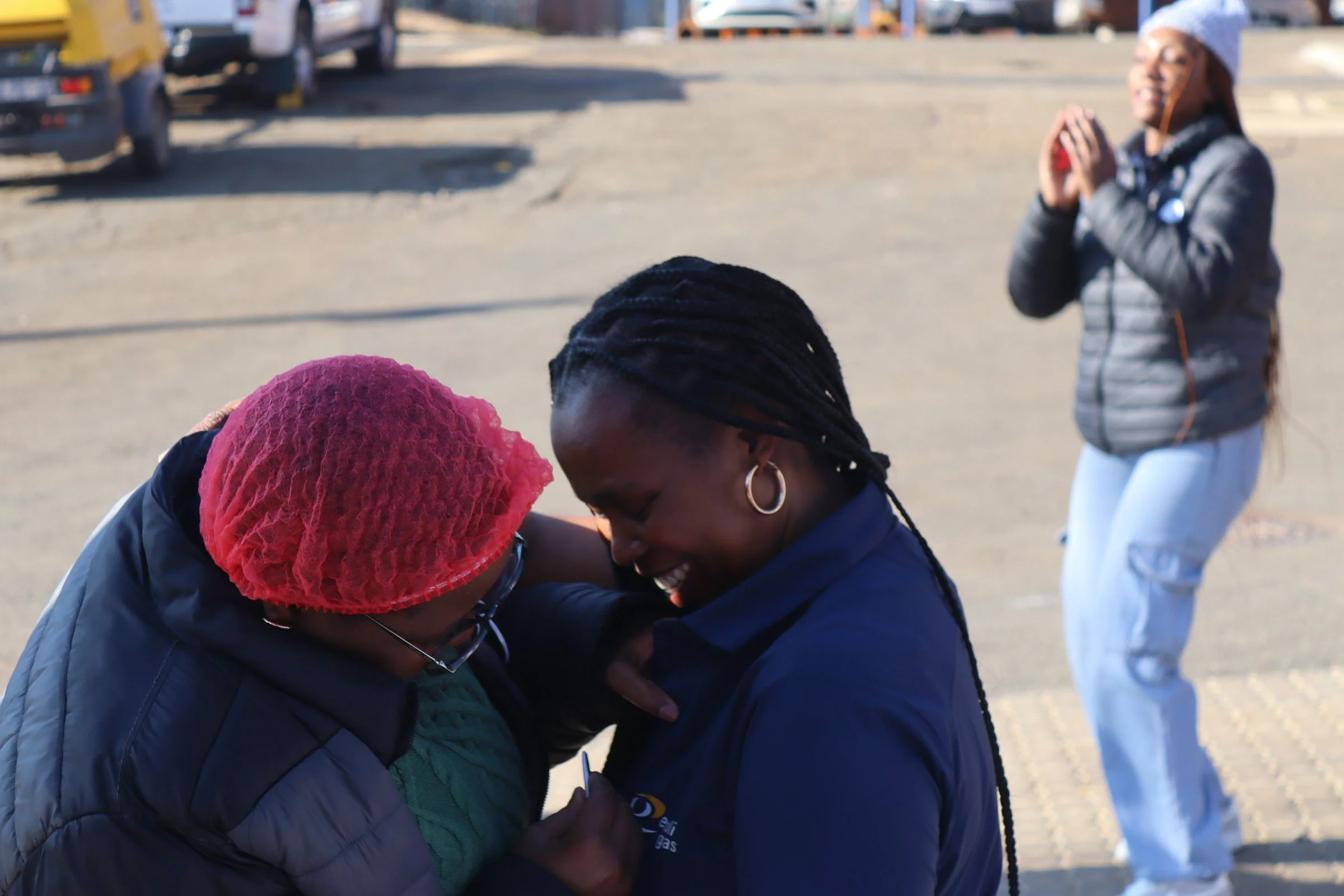 Three people are hugging and smiling outdoors in a parking lot, with a woman standing in the background clapping and smiling.