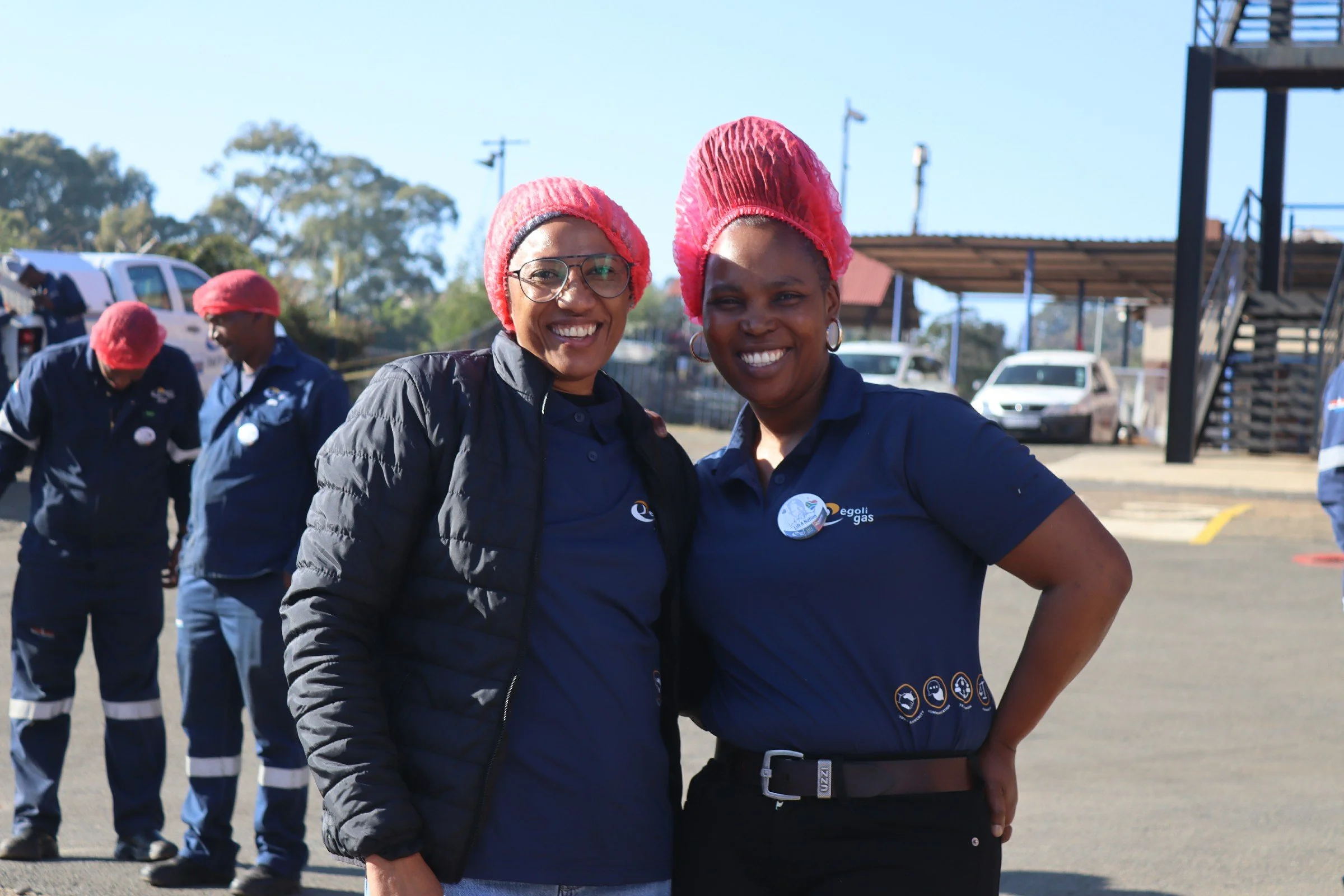 Two smiling women wearing pink hair nets and dark blue uniforms standing outdoors, with other workers in similar uniforms in the background.