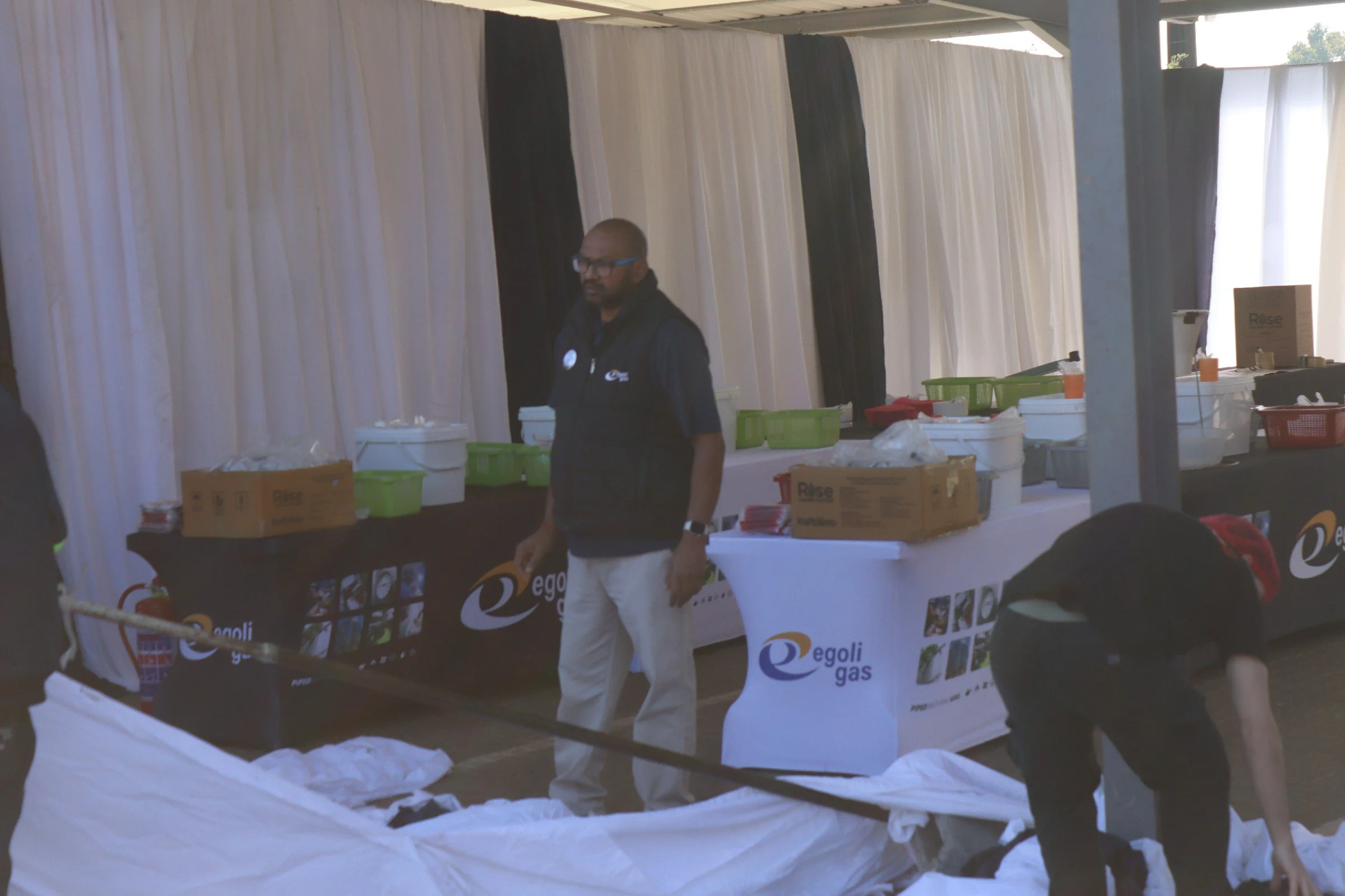 A person wearing glasses and a black vest standing behind a table with Egoli Gas promotional materials at an indoor event. The table is covered with white cloth and has boxes, green baskets, and flyers. Behind is a backdrop of white and black curtain