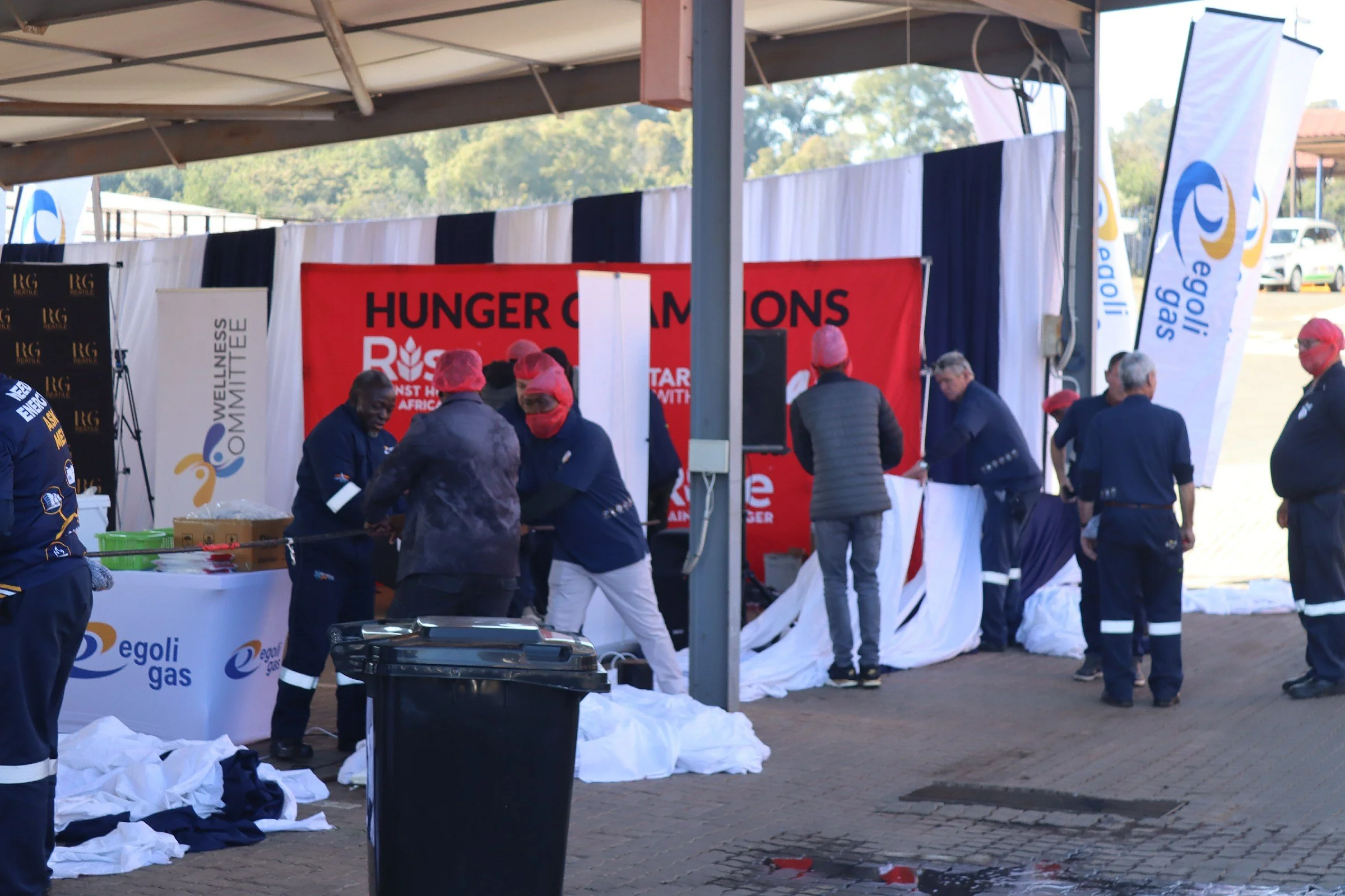 People in protective clothing and masks preparing for a humanitarian event at an outdoor venue with banners for Hunger Champions and Egoli Gas.