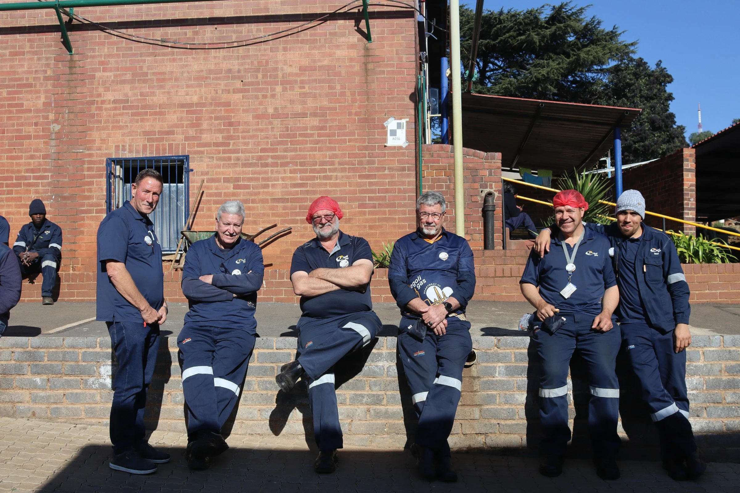 Group of seven people wearing dark blue uniforms, standing and kneeling on steps outside a brick building with a blue window and trees in the background. Some are smiling, and one person is wearing a red headscarf.