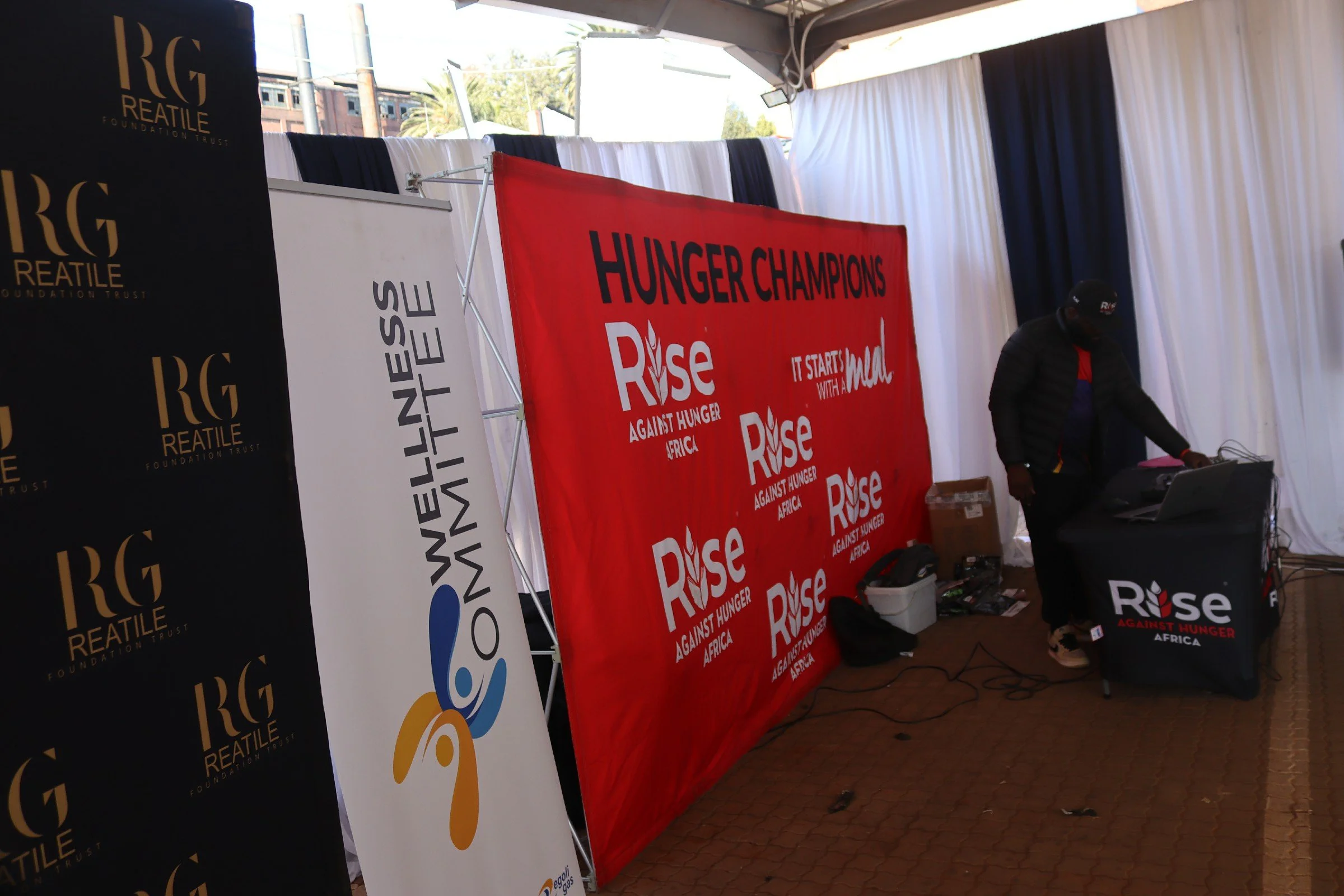 A display setup for a hunger relief event with banners that read 'Raise Against Hunger Africa' and 'Hunger Champions'. There is a man working with electronic equipment at a table, and the area is covered with draped fabric walls and banners.