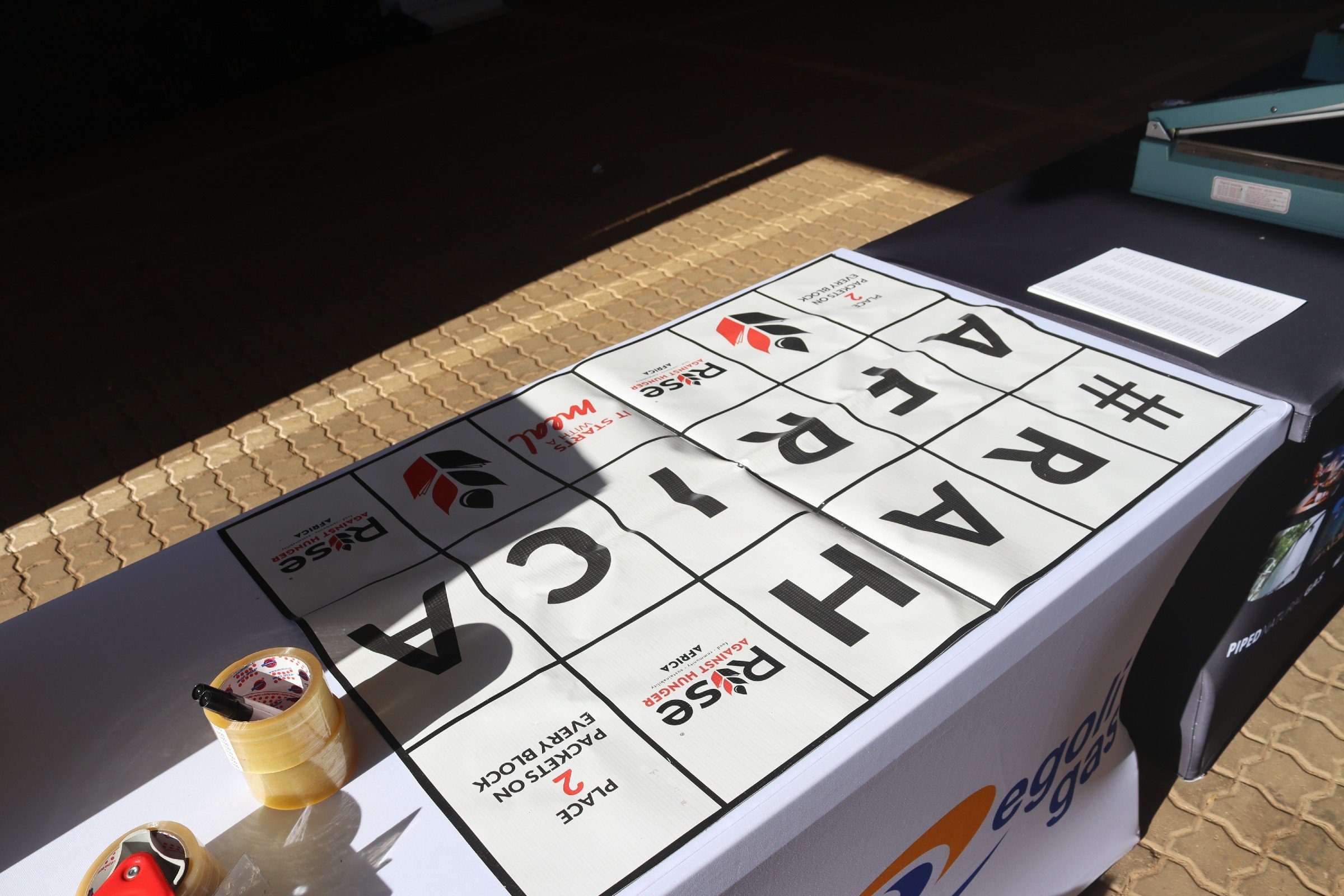 Table with signs for a racing event, marked with the brand 'DAS Racing' and labeled as 'Race on Picket Baby Black.' There are small rolls of tape, a black marker, and printed sheets on the table, and the scene is partially illuminated with sunlight.