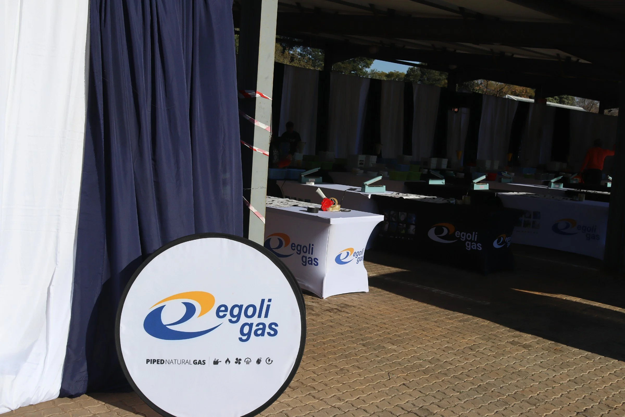 An outdoor market booth with Egoli Gas branding, featuring a table with promotional materials and a backdrop with curtains, set on a brick paved area under a shelter.