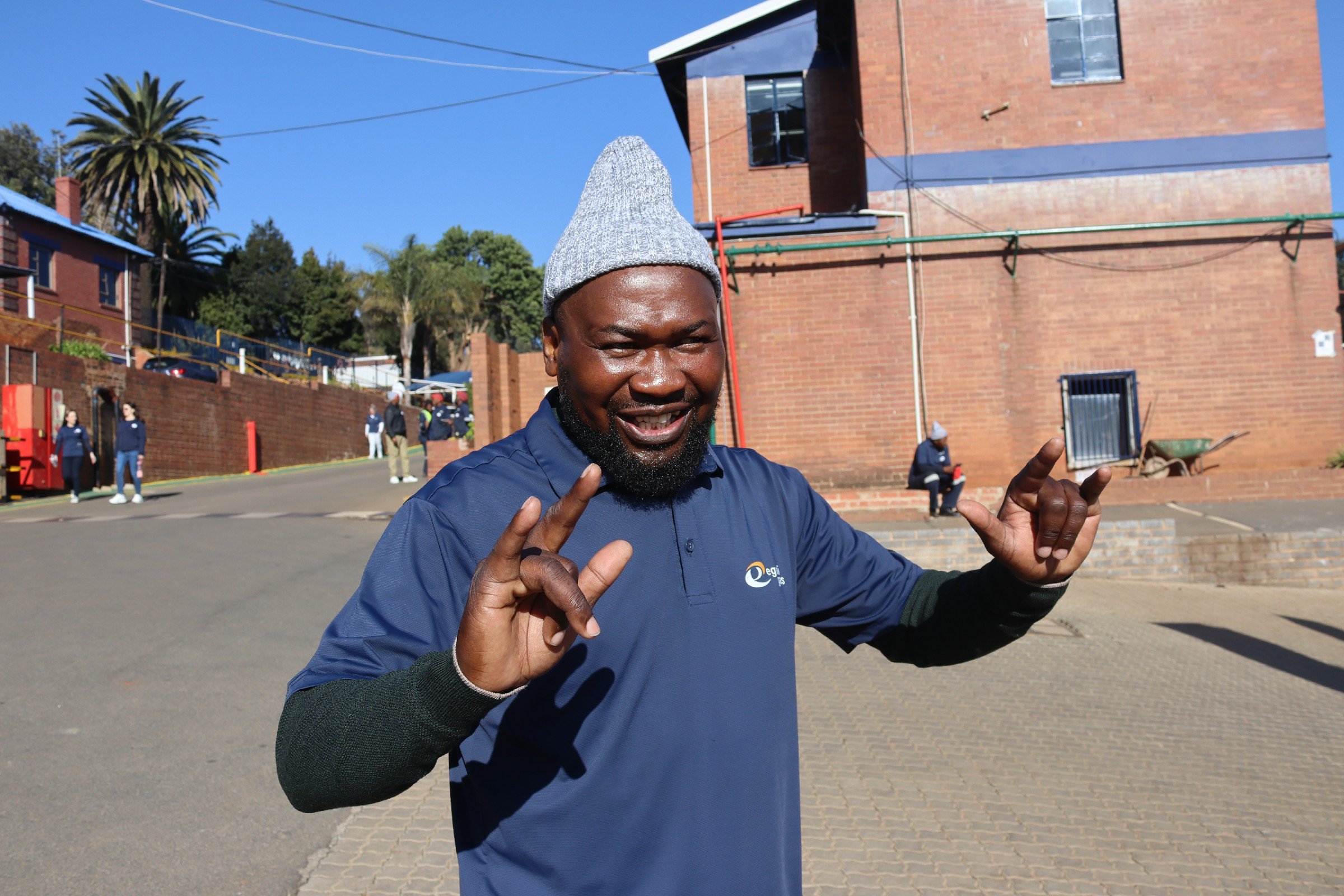 A man wearing a gray knit cap and a blue long-sleeve shirt is smiling and making hand gestures outdoors, with a brick building, trees, and a clear blue sky in the background.