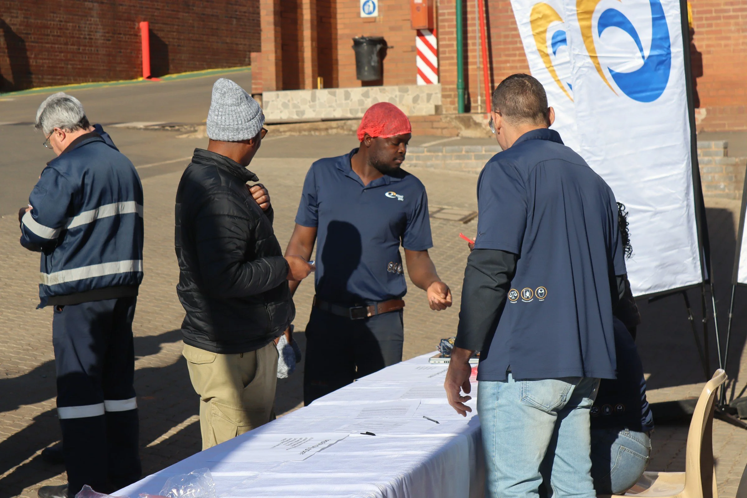 Group of people gathered around a table outside, engaged in conversation or paperwork, with a brick building and a flag in the background.