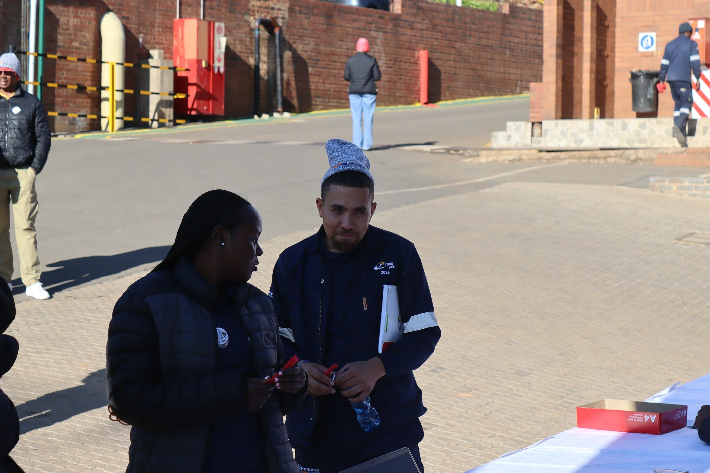 Two women and a man are gathered around a table outdoors, with others walking in the background. The women are focused on a phone, and the man is holding a pen. People are dressed warmly, and the scene appears to be during daytime.