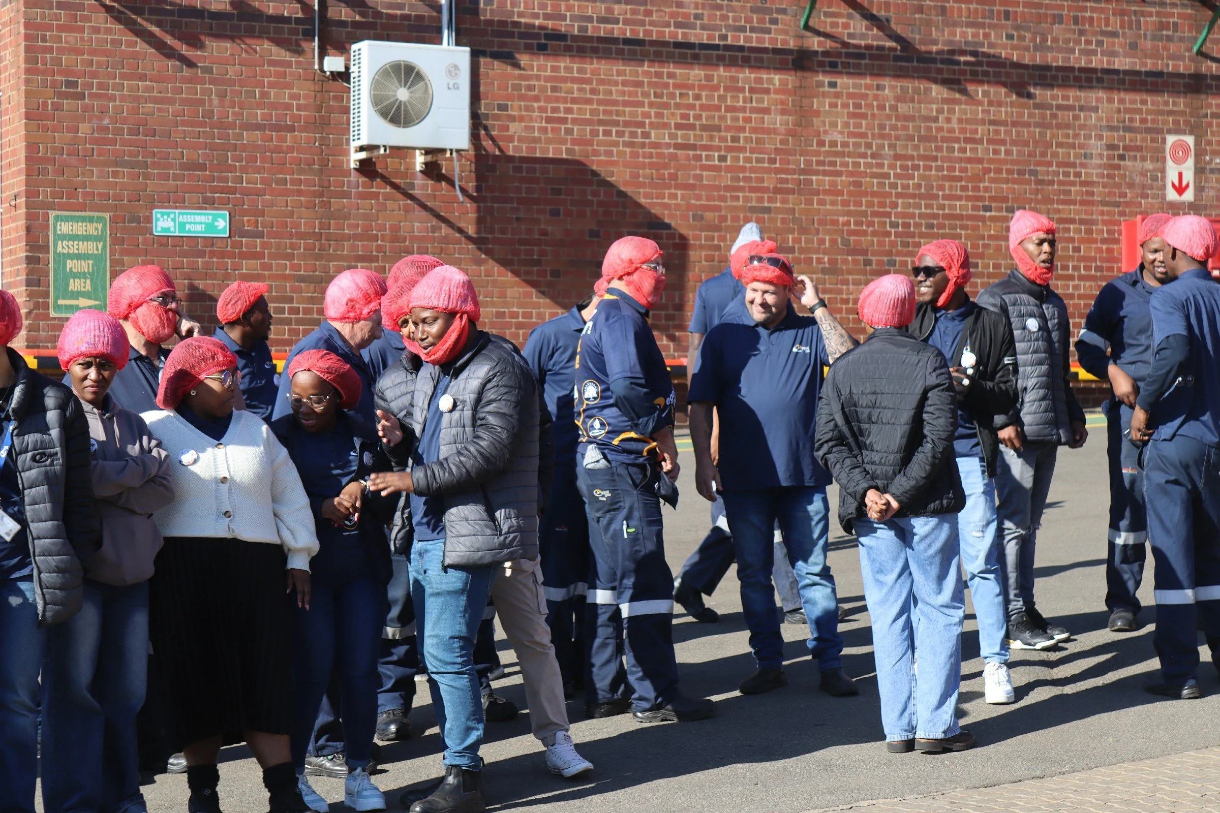 Group of people wearing red hairnets and casual clothing, standing outside near a brick wall with safety signs.
