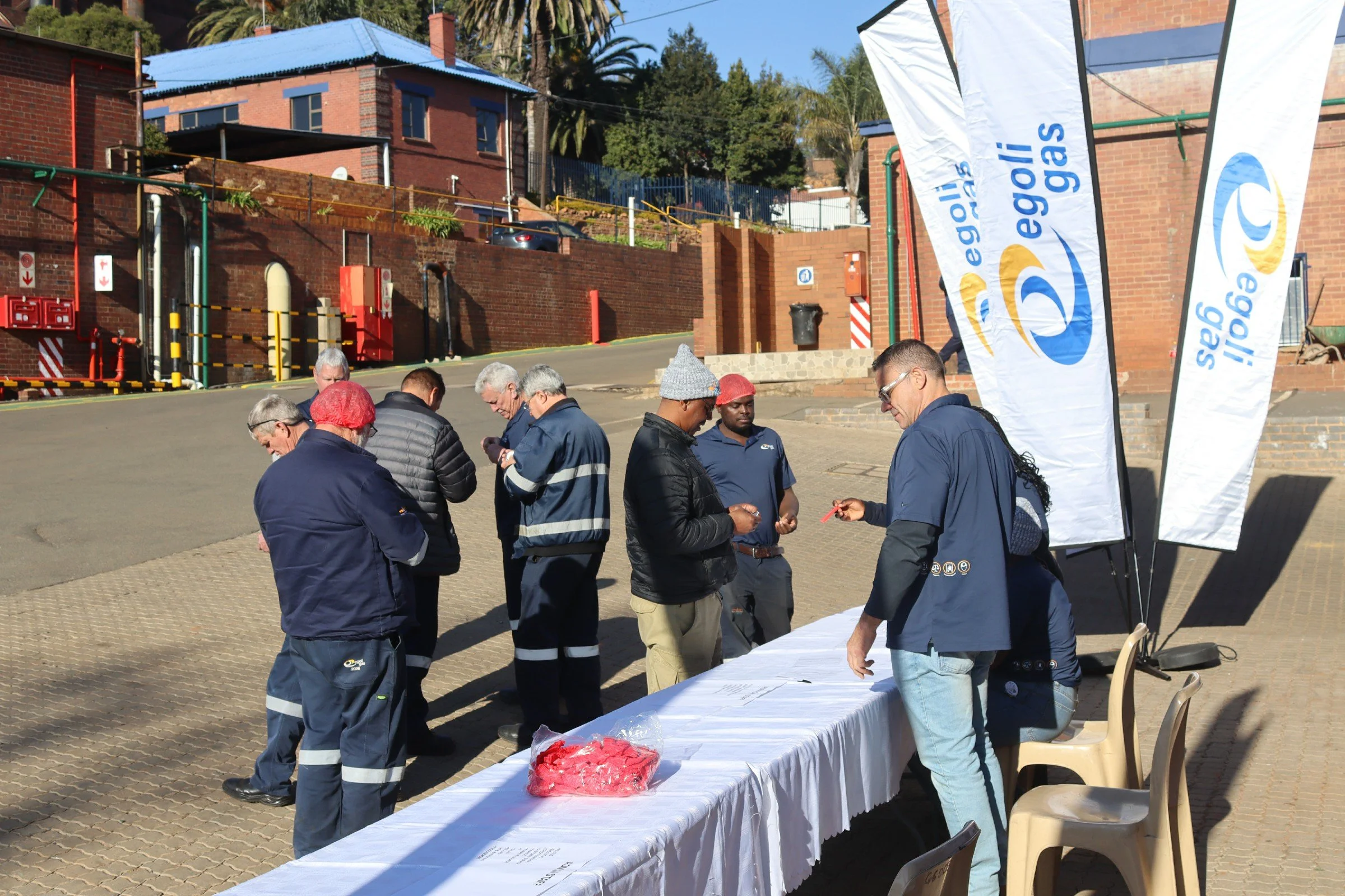 Group of people gathered around a table with papers and a box of heart-shaped candies, outside next to flags that say 'Egglisi Eggas' on a sunny day.