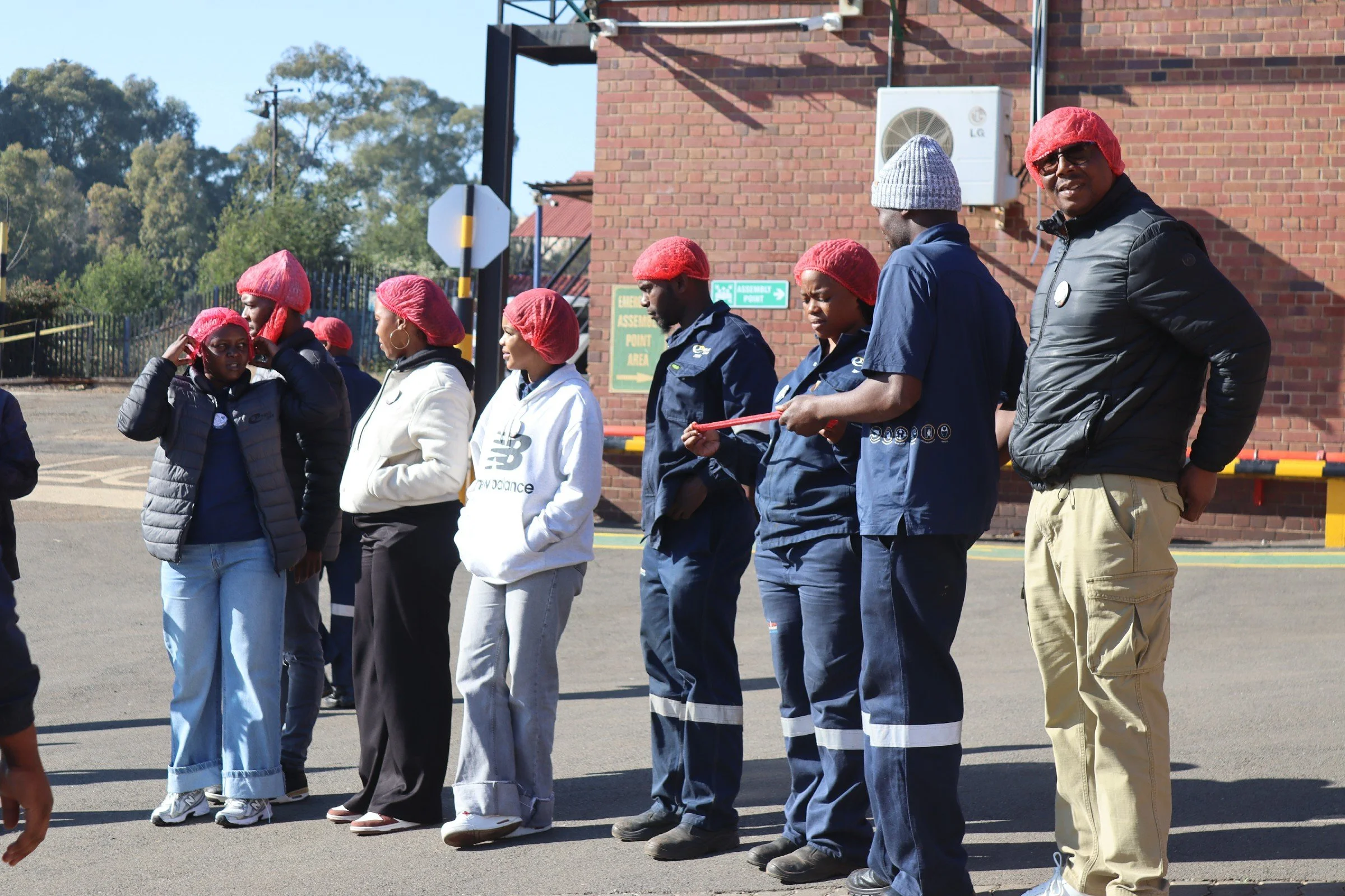 Group of people standing in line outdoors, wearing red hair coverings and casual clothing, with a brick building and trees in the background.